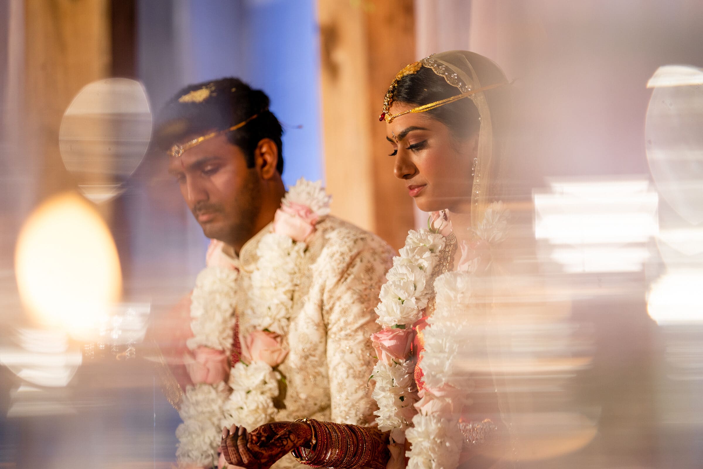 A bride and groom sit together during a traditional wedding ceremony. They are adorned in ornate attire and garlands. The lighting is soft and warm, adding a dreamy effect to the scene.