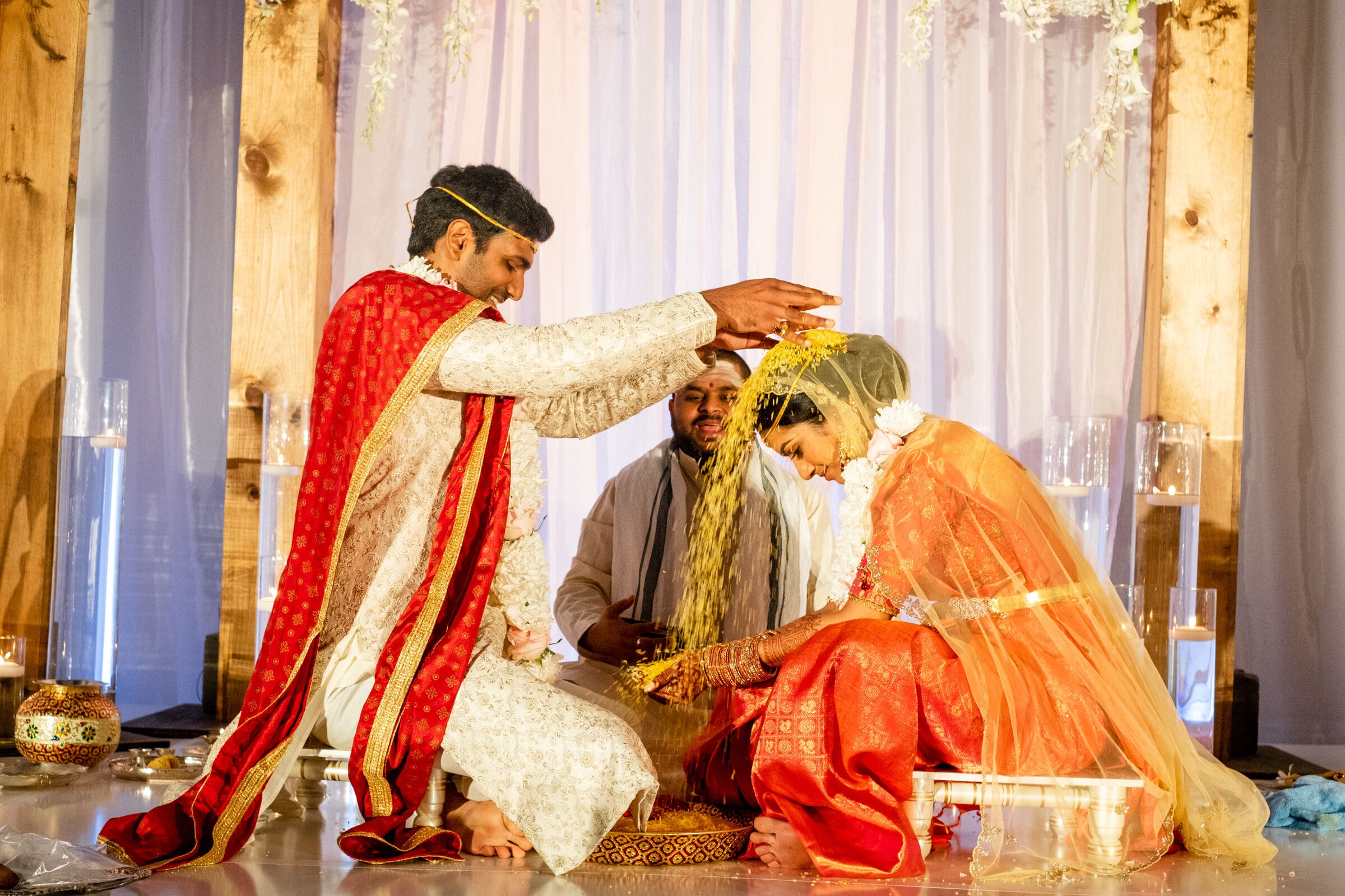 A man in traditional attire pours grains over a seated woman wearing an orange saree and veil. They are participating in a cultural ceremony, surrounded by candles and fabric drapes, as an officiant observes.