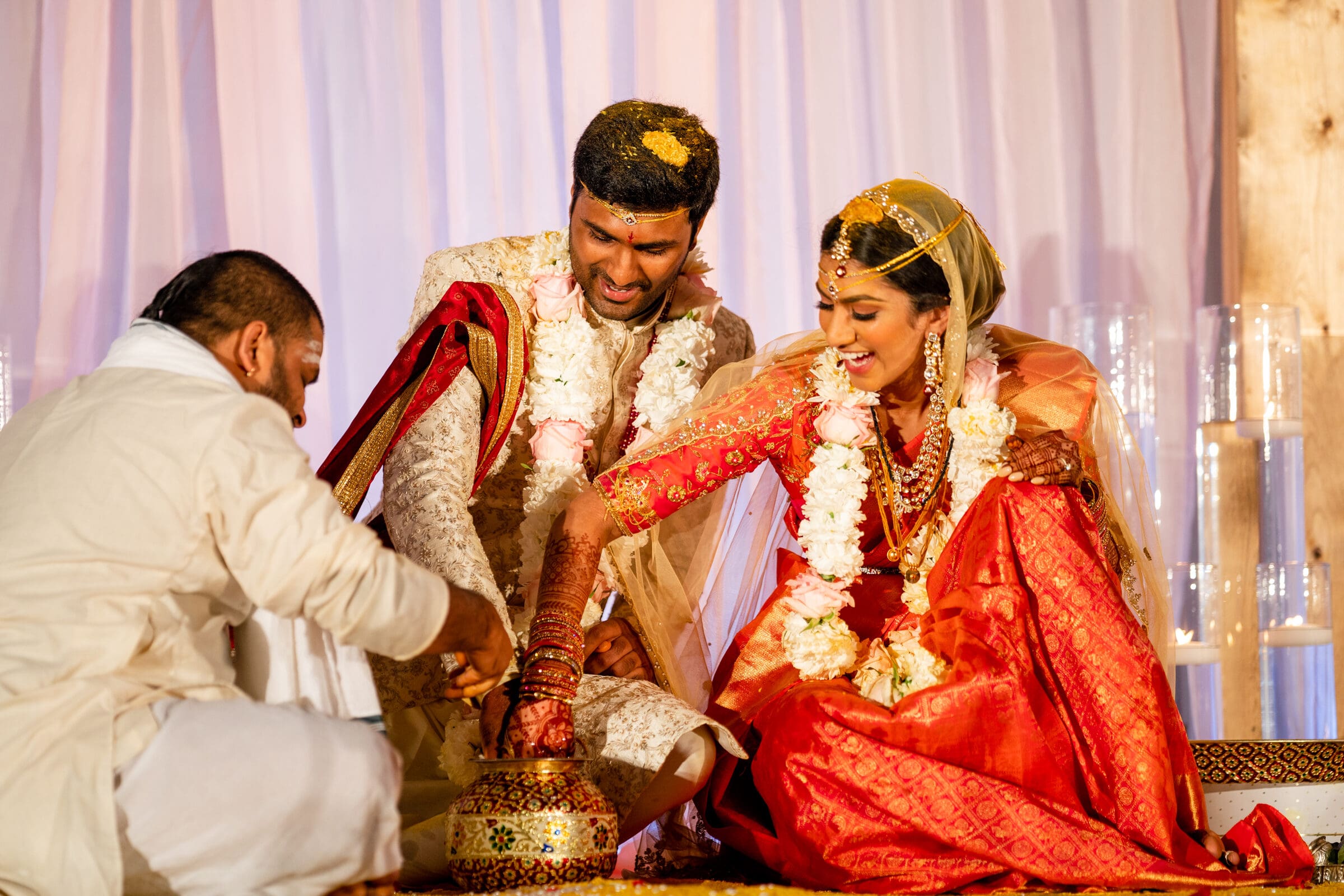 A couple dressed in traditional Indian attire participates in a wedding ceremony. They are adorned with floral garlands and seated, while a priest assists with rituals. The bride wears a red sari; both are smiling under a decorative backdrop.