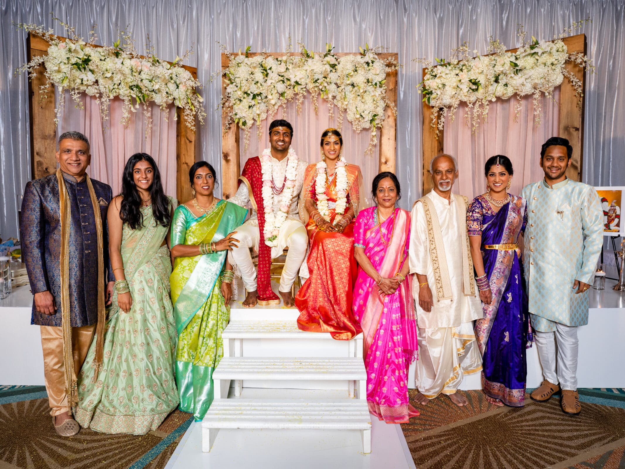 A joyful wedding scene with a couple in traditional attire seated on a flower-adorned stage. Theyre surrounded by eight family members, all wearing colorful, elegant clothes, smiling happily. The background features decorative floral arrangements.