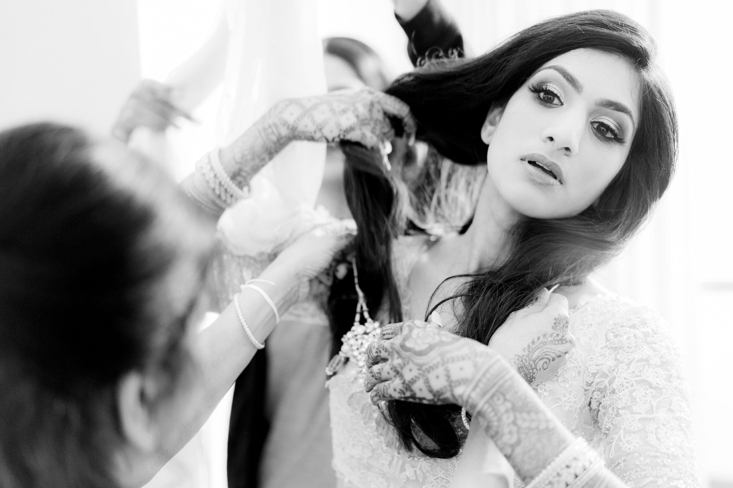 A black and white photo of a bride with intricate henna on her arms getting ready. She holds her long hair as attendants, partially visible, assist her. Her expression is focused, wearing an ornate lace outfit.