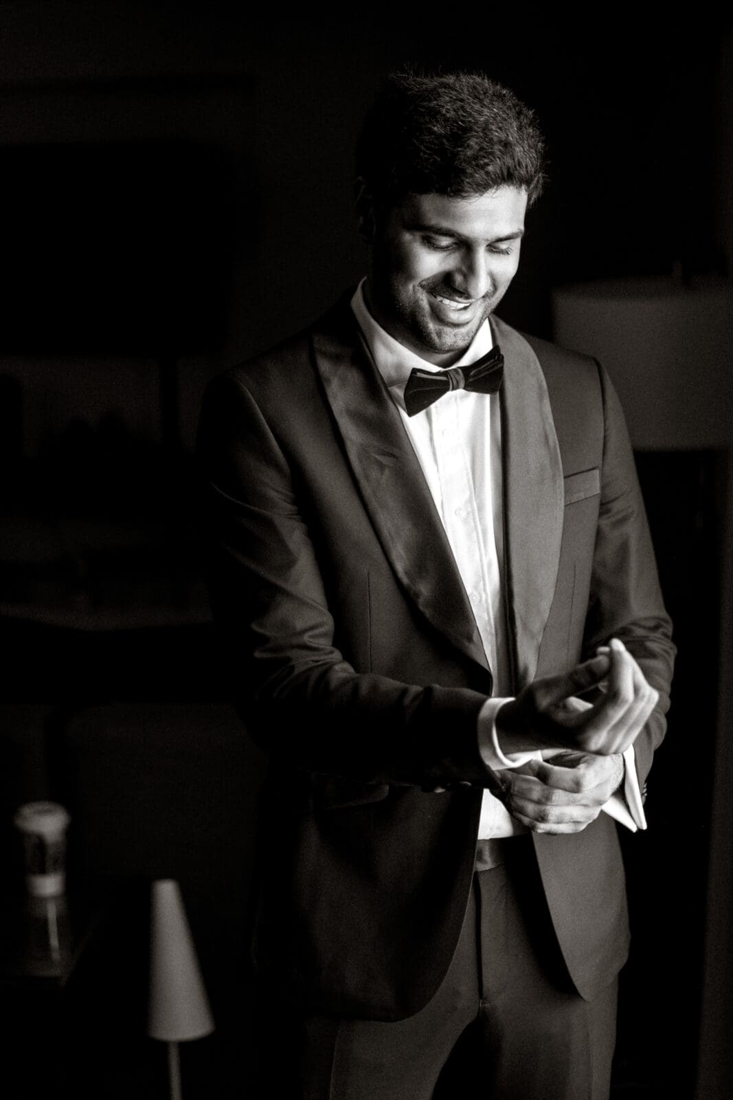 A man in a tuxedo adjusts his cufflink, smiling slightly. The setting is dimly lit, creating a classic, elegant atmosphere. A small table with a cup is visible in the background. The photo is in black and white.