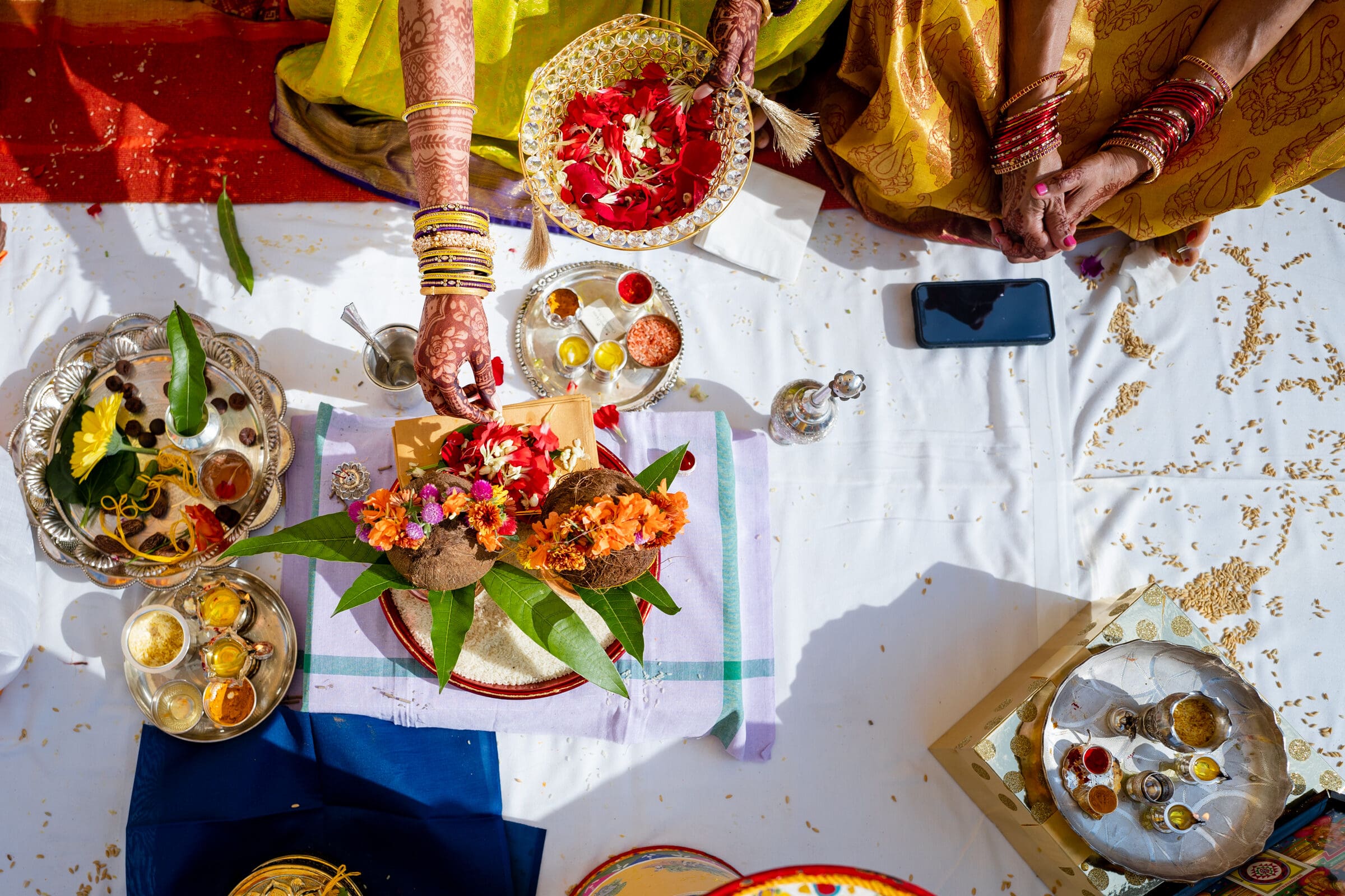 A vibrant Hindu ceremony setup with colorful flowers, coconut, and leaves on a cloth. Women in traditional attire with bangles are arranging items. Assorted bowls with spices and a smartphone are visible, capturing the festive atmosphere.