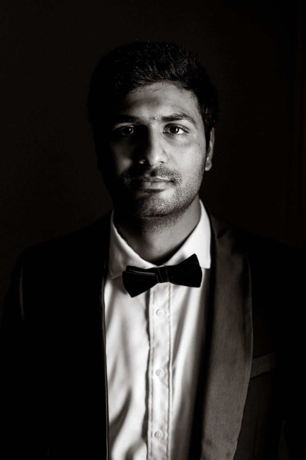 A man in a tuxedo with a bow tie looks directly at the camera. The image is in black and white, with dramatic lighting highlighting one side of his face, creating a classic and sophisticated atmosphere.