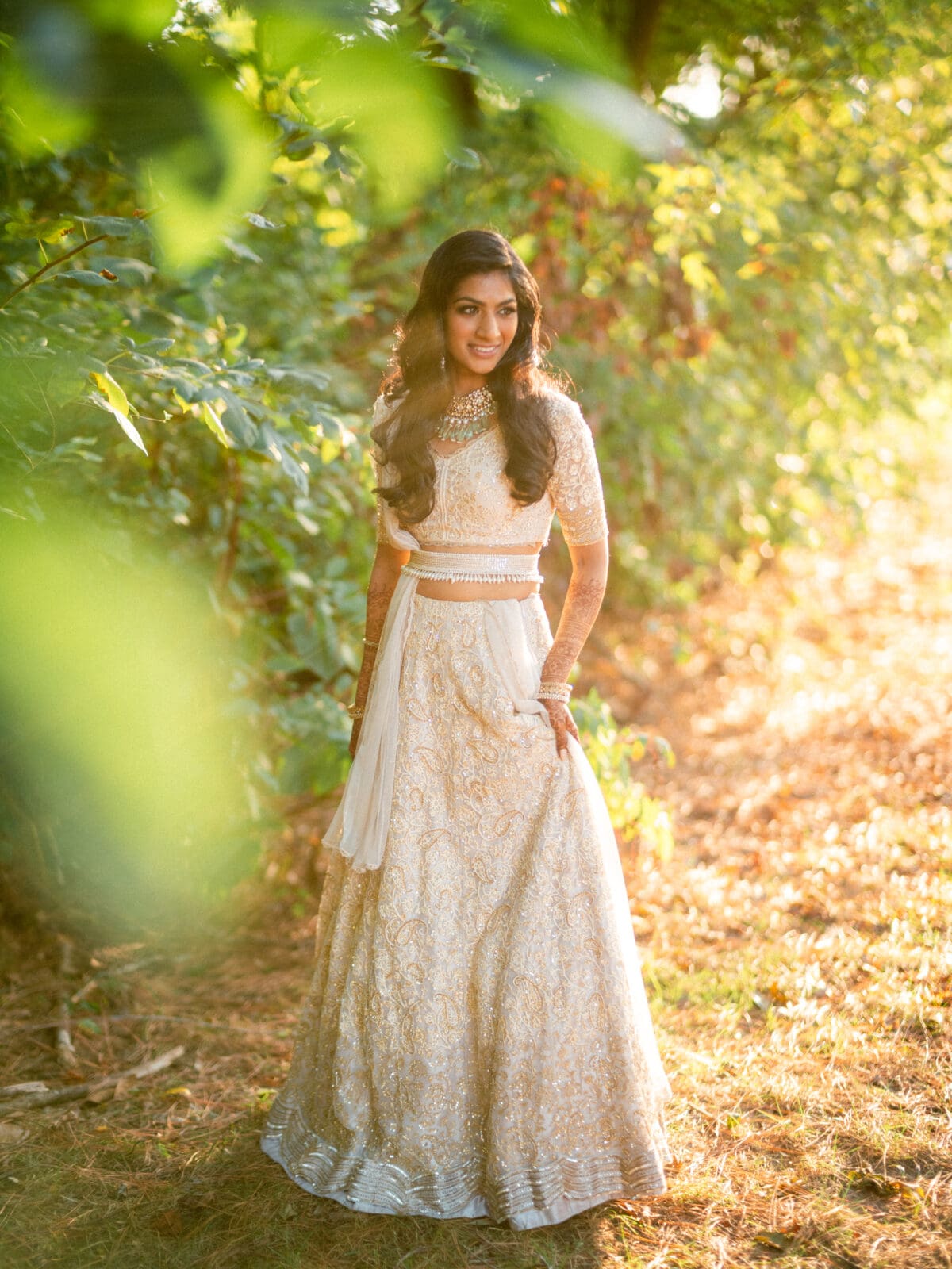 A woman in a cream-colored, intricately embroidered dress stands outdoors among lush greenery. She smiles softly in the warm sunlight filtering through the leaves, creating a serene and elegant atmosphere.