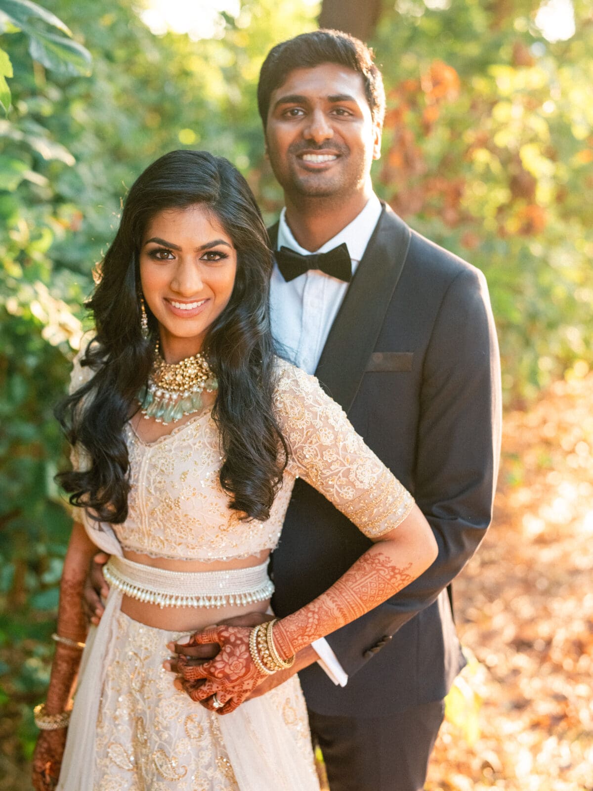 A couple dressed in formal attire stands outdoors. The woman is wearing a light-colored, embellished outfit with henna on her hands, while the man is in a dark suit with a bow tie. They are smiling, surrounded by greenery and sunlight filtering through.