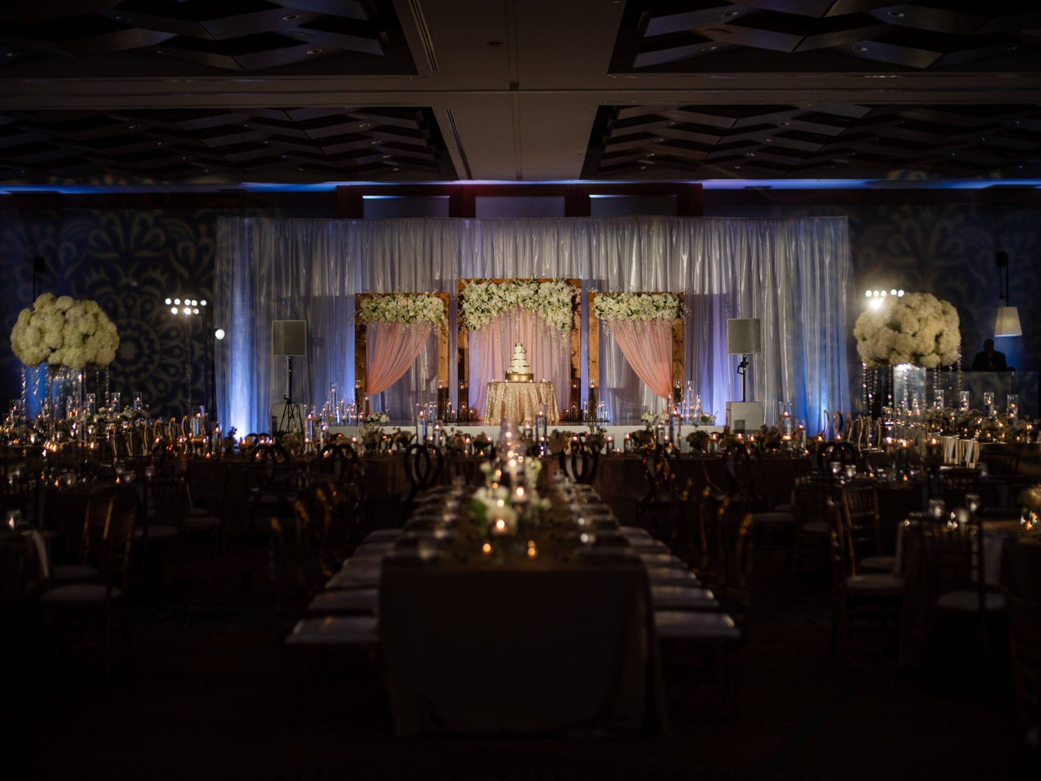 A dimly lit wedding reception hall features elegantly set tables with candles and floral centerpieces. At the center, a tiered wedding cake is displayed on a stage draped with curtains and adorned with flower arrangements.