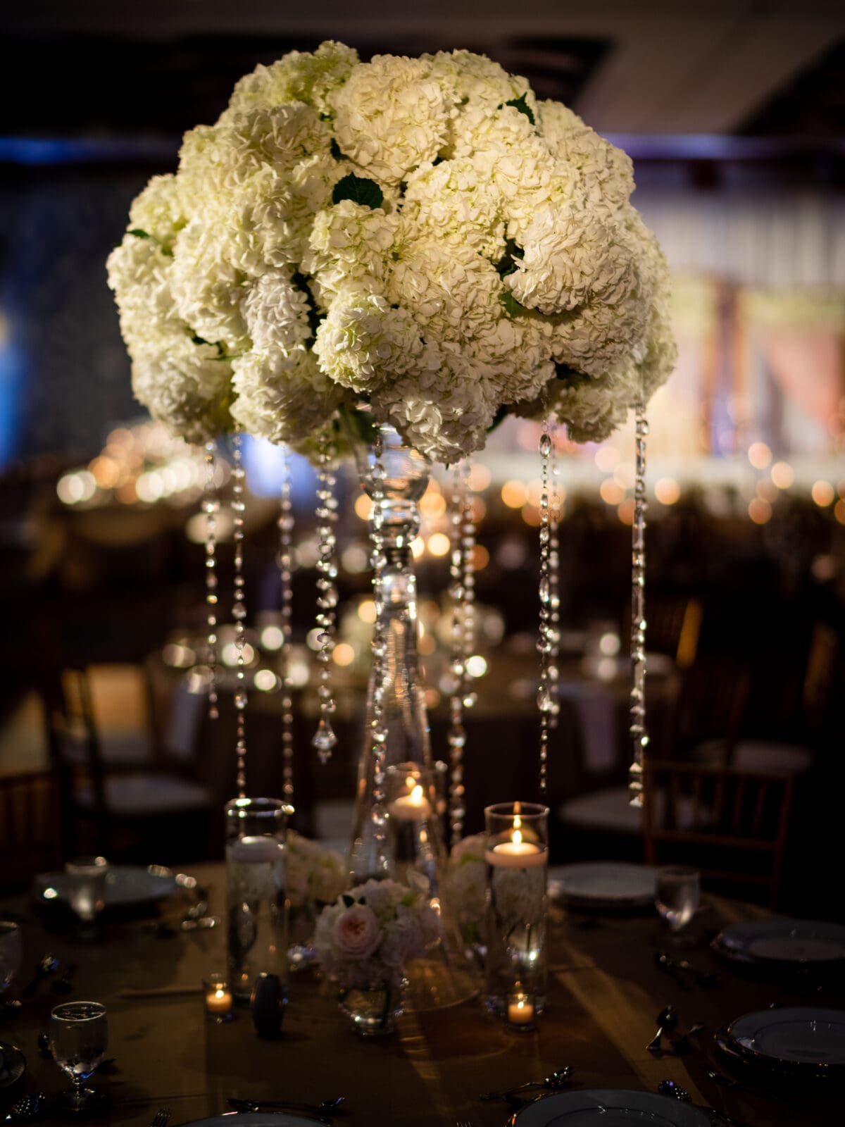 A tall, elegant centerpiece with white hydrangeas in a clear vase adorned with hanging crystal strands. It is surrounded by lit candles on a round dining table, creating a warm, romantic atmosphere.