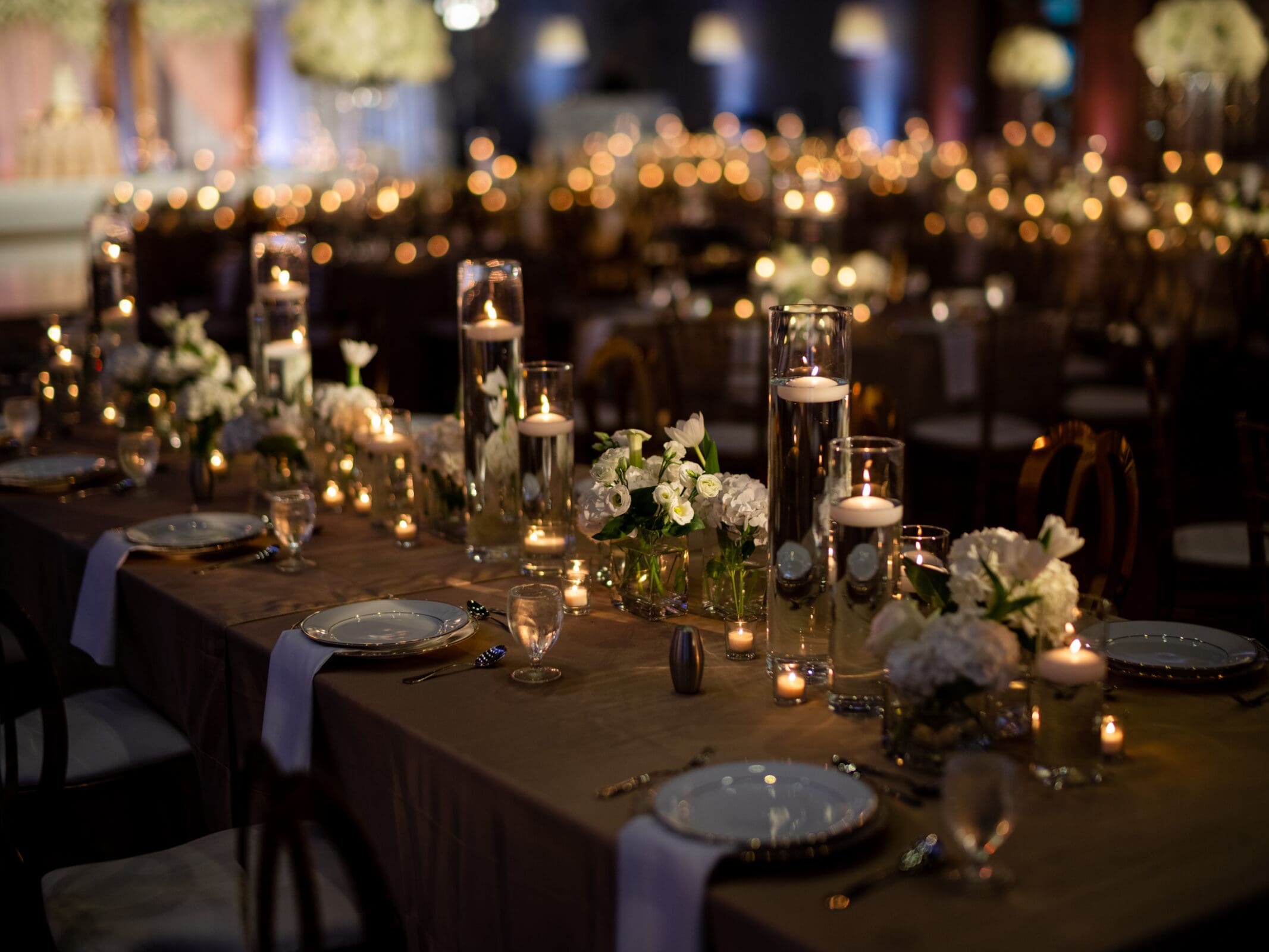 A dimly lit table is elegantly set for a formal event. It features white flowers, glass candle holders with lit candles, and white plates with silver cutlery. The background is softly illuminated, adding a warm ambiance.
