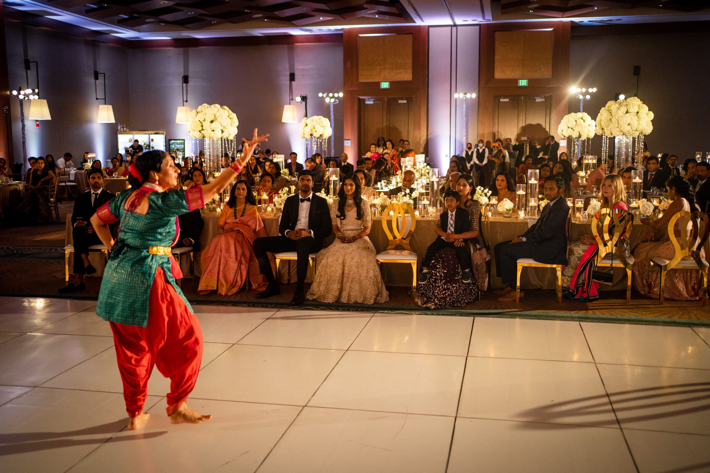 A performer in traditional attire dances on a white stage, captivating a well-dressed audience sitting at tables in a warmly lit banquet hall decorated with tall floral centerpieces.