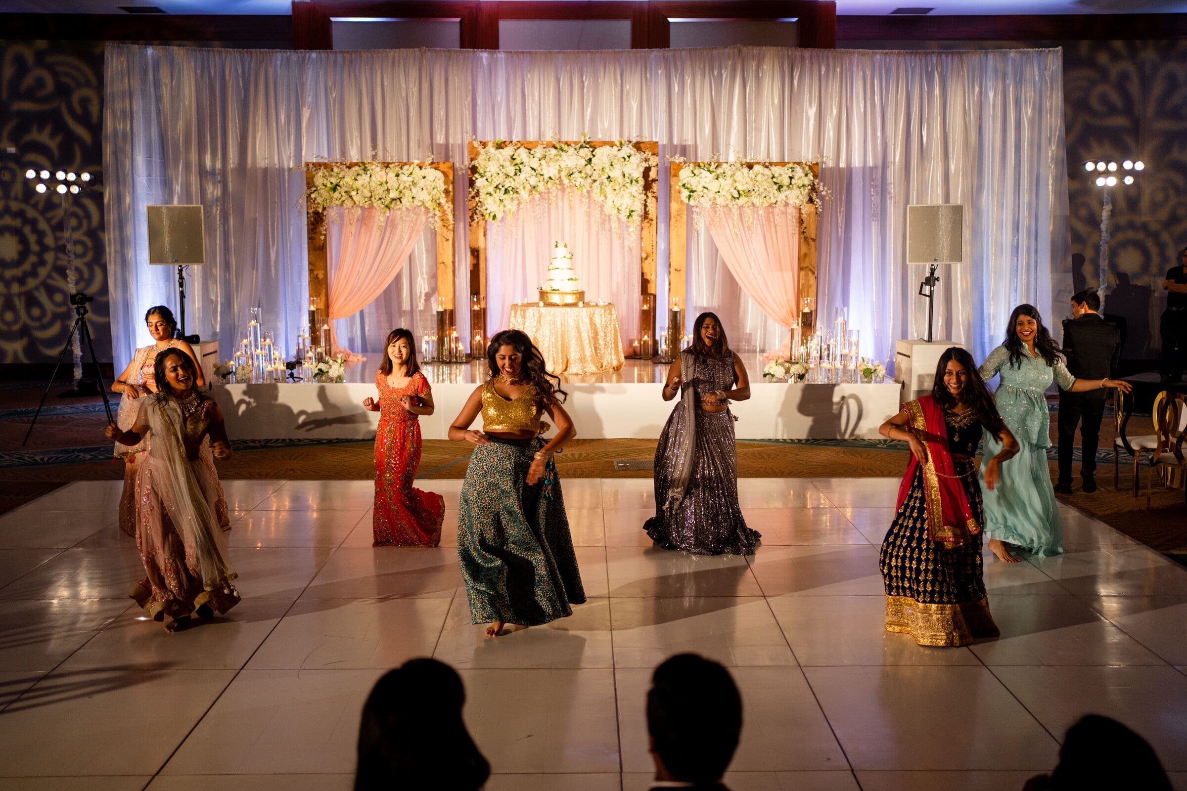 A group of women in colorful traditional attire perform a choreographed dance at an indoor event. In the background, a stage is decorated with flowers, curtains, and a tiered cake. The lighting is warm and festive.