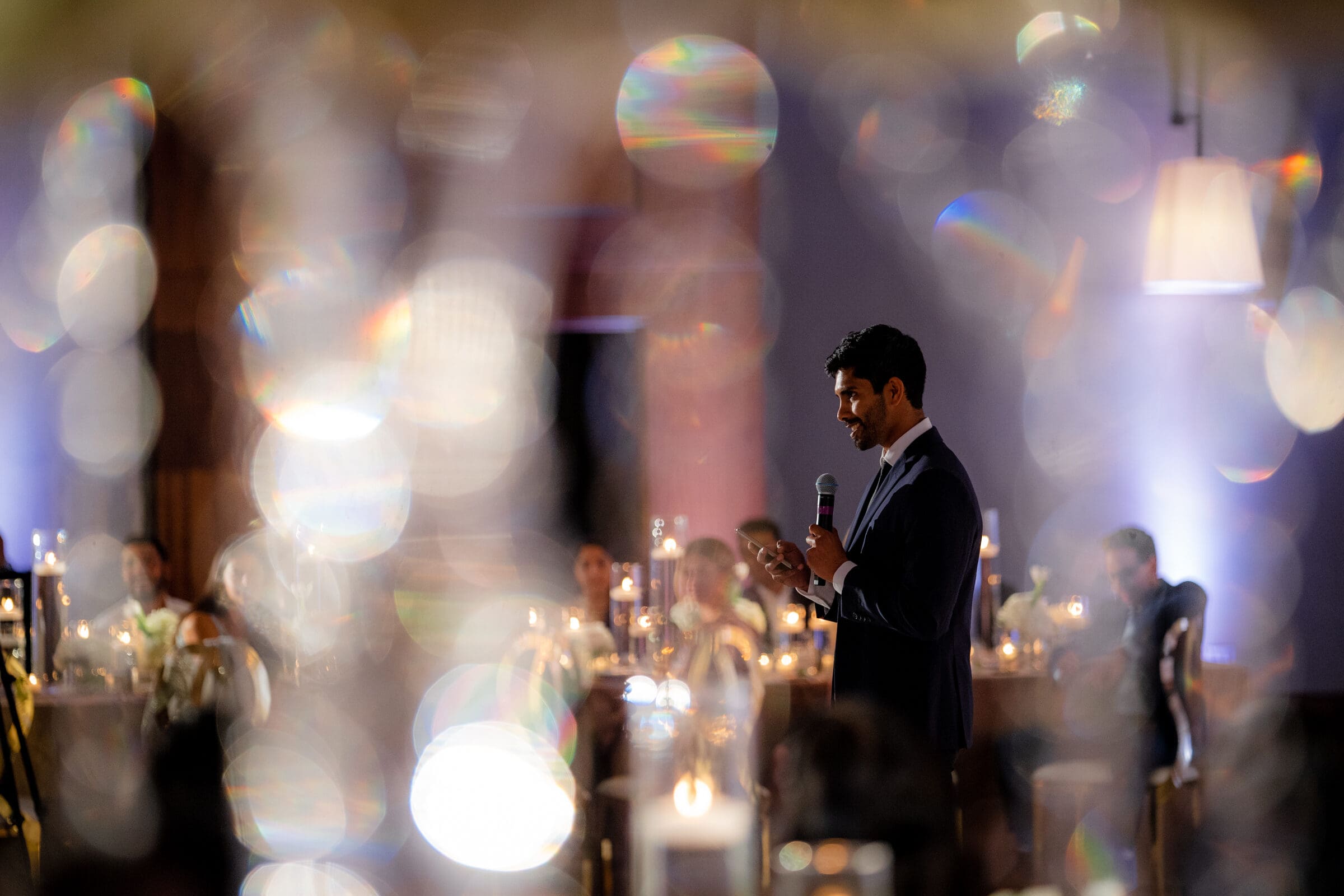 A person in a suit speaks into a microphone at an event. The background is blurred with colorful, circular lights, creating a dreamy effect. Candles and softly lit tables are visible in the room.