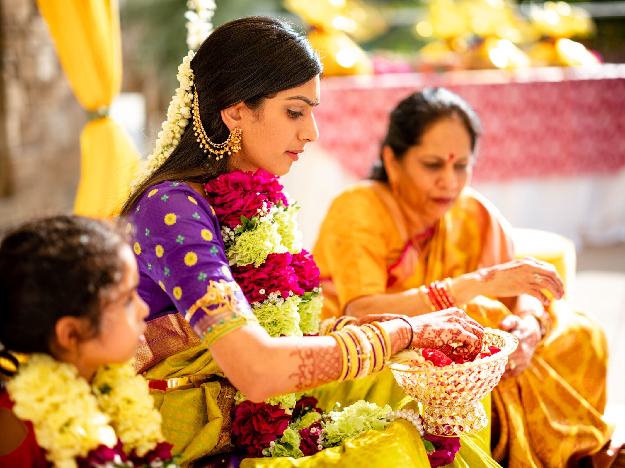 A woman in a purple and gold sari with floral garlands participates in a traditional ceremony with an older woman in a yellow sari. A child sits nearby, also adorned with flowers. The setting is festive with bright colors.