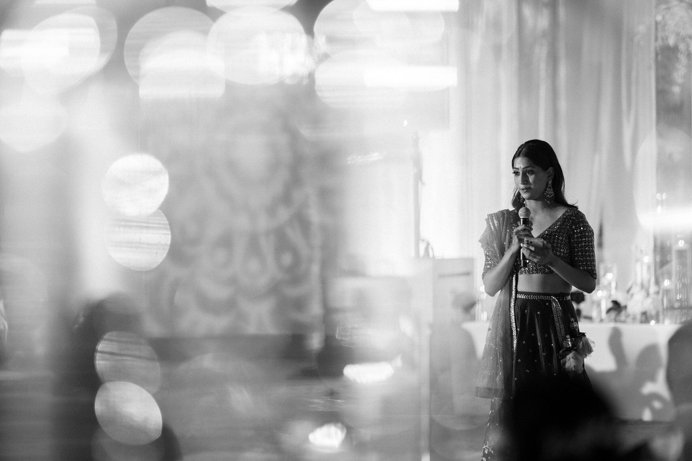 A woman in traditional attire stands on stage holding a microphone, set against a softly blurred, illuminated background. The image conveys a dreamy, ethereal atmosphere with delicate light bokeh effects surrounding her.