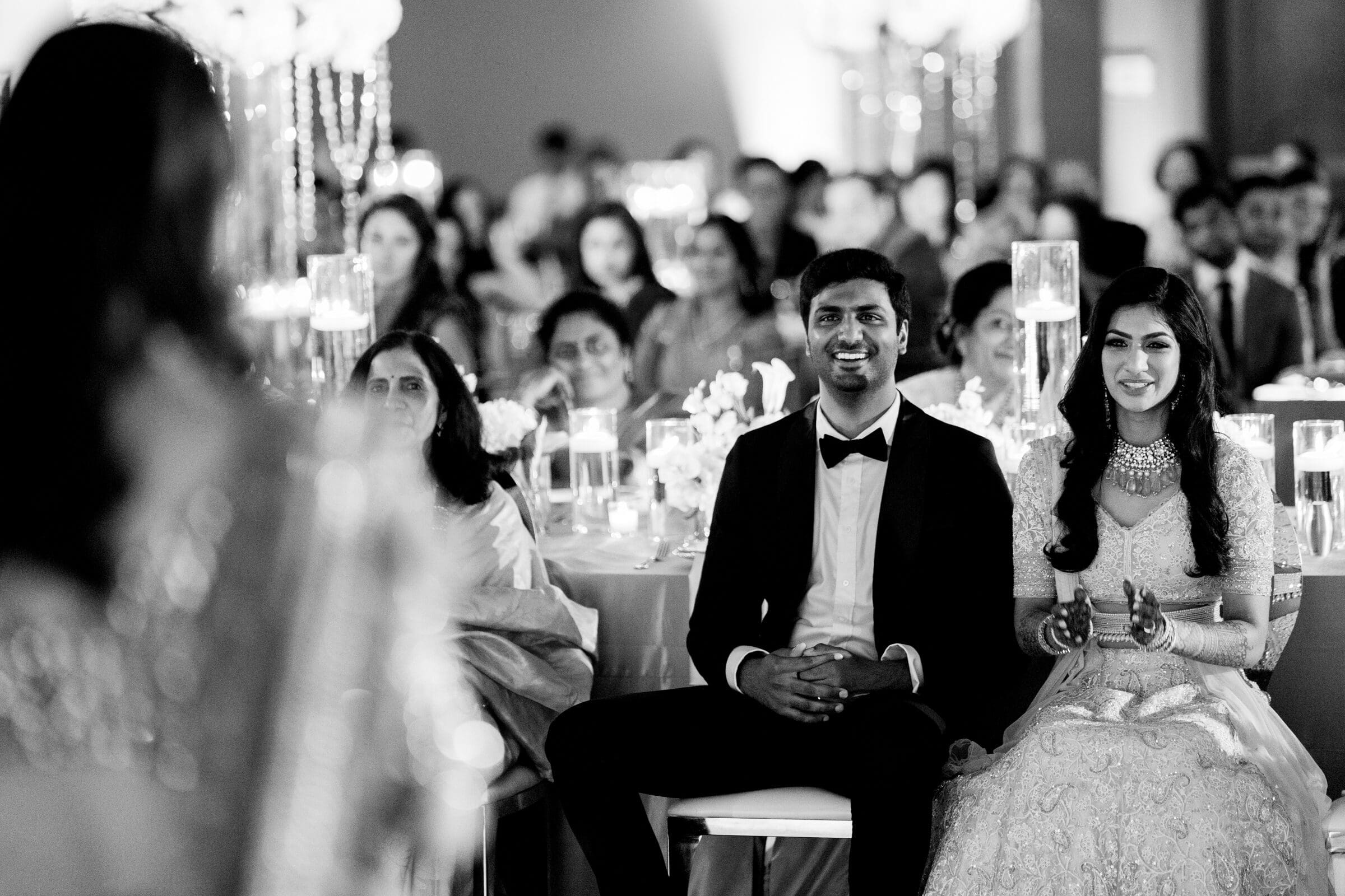 A black and white photo shows a seated couple at a formal event. The man, in a suit, and the woman, in an embellished gown, smile and clap. They are surrounded by an attentive audience in an elegantly decorated setting.