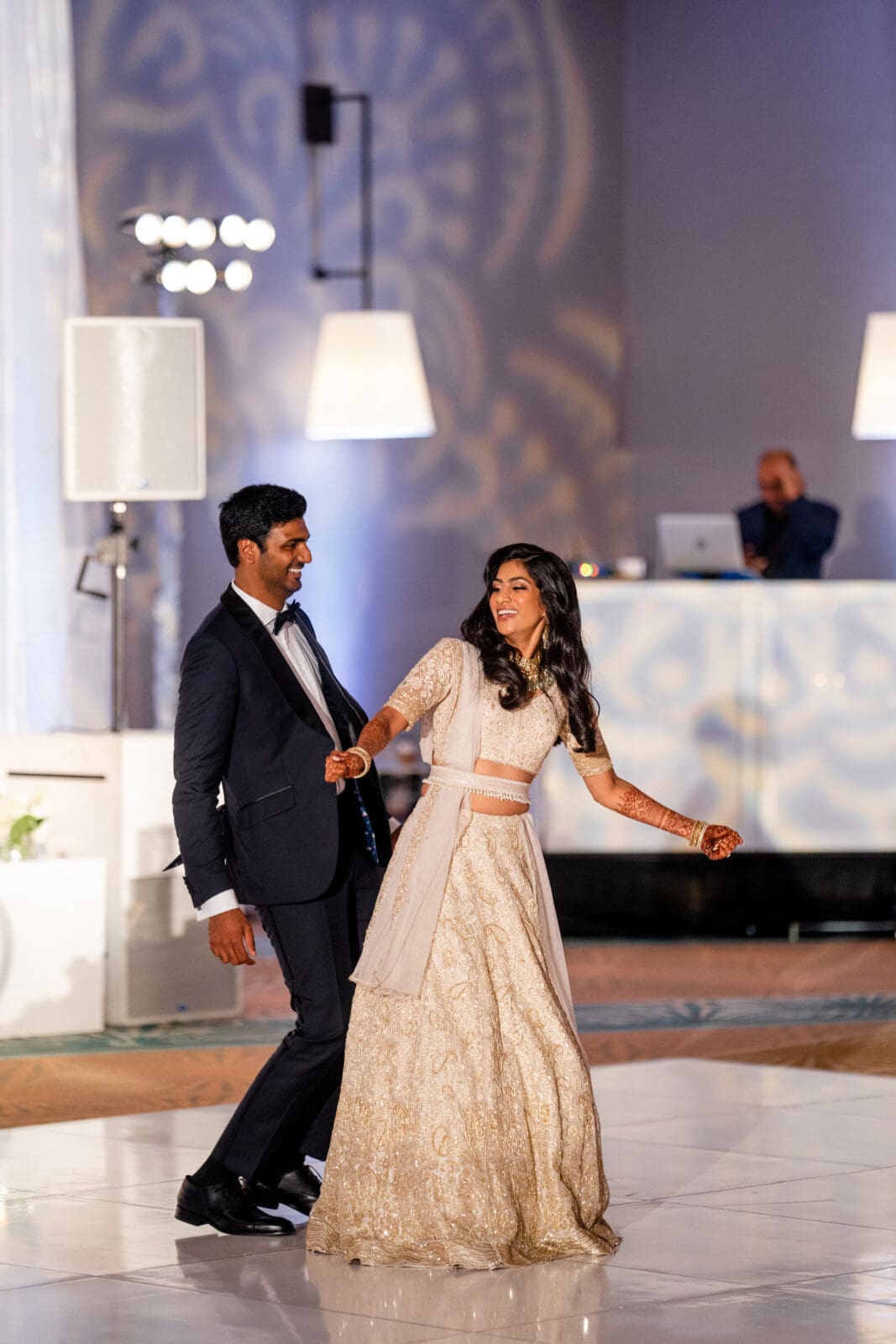 A couple dances joyfully at an indoor event. The man wears a dark suit, and the woman is in a light-colored traditional dress. The background features decorative lighting and a DJ booth with a person standing behind it.