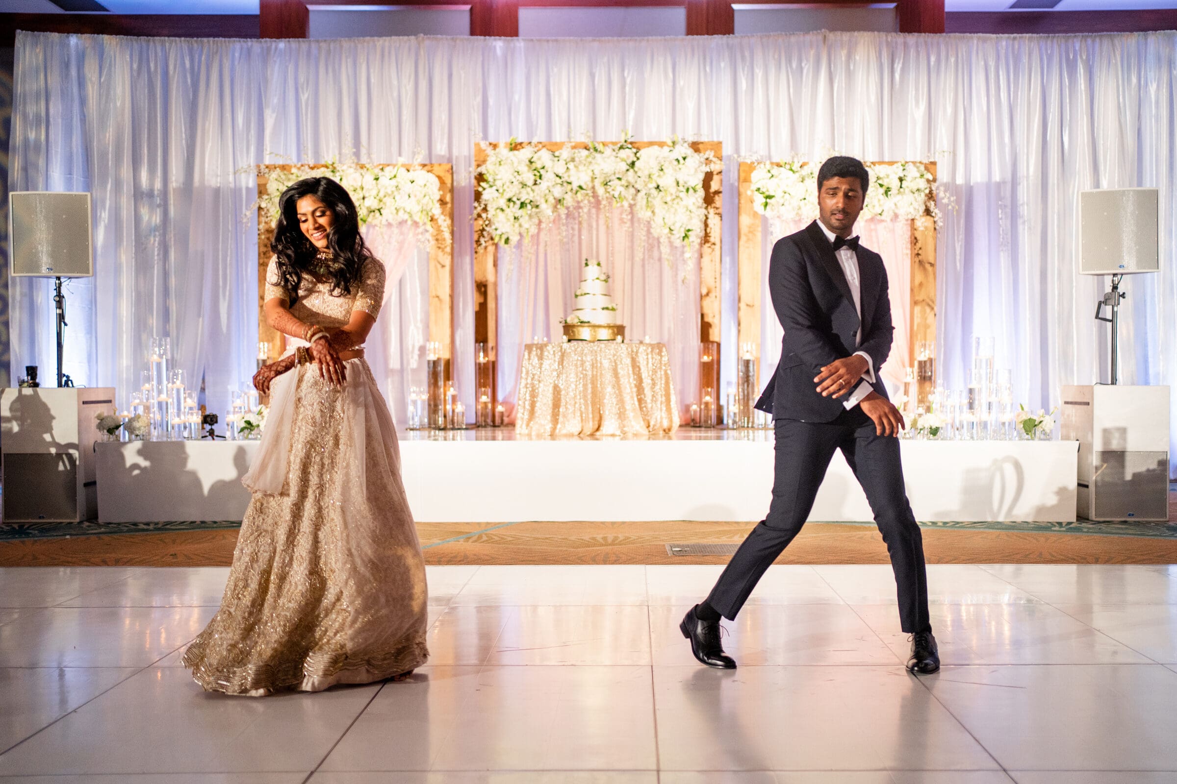 A couple dances on a polished floor in a decorated hall. The woman wears a flowing white gown, while the man is in a dark suit. Behind them is a tiered cake and a backdrop with white and gold flowers. The setting is elegant and festive.