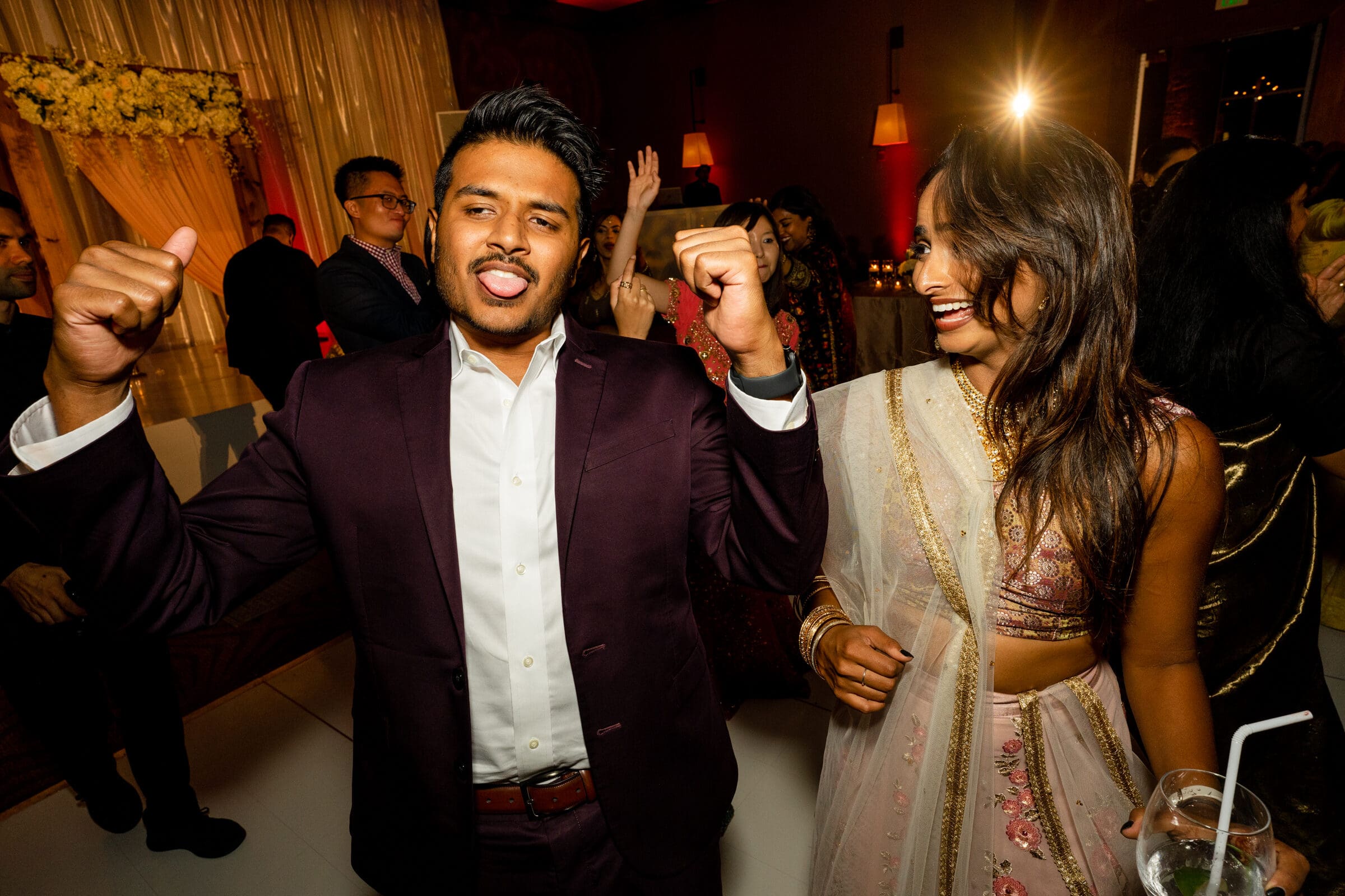 A man in a dark suit enthusiastically poses with his arms bent upwards, while a woman in a colorful sari smiles beside him at a lively indoor gathering. Guests and warm lighting create a festive atmosphere in the background.