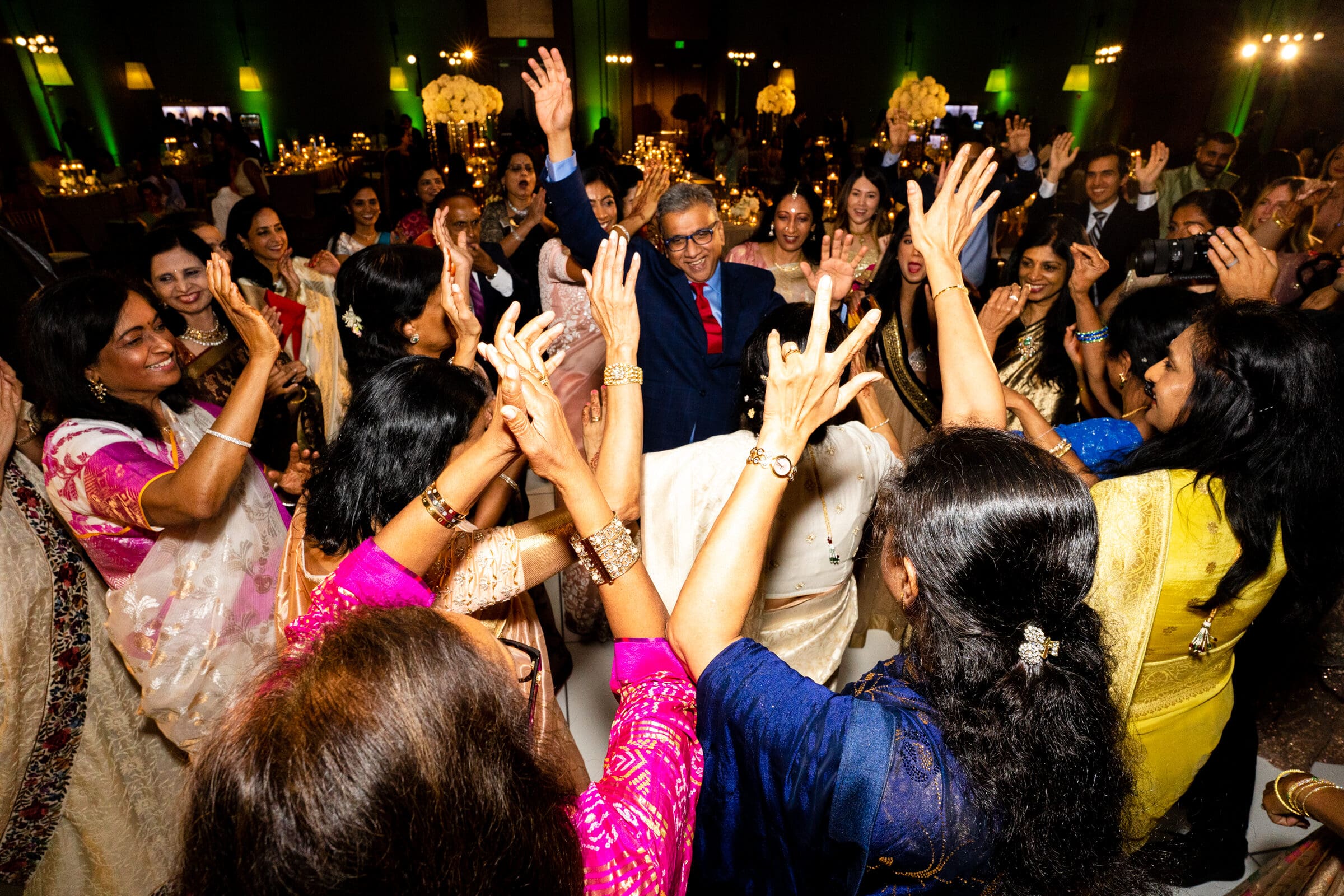 A large group of people in colorful traditional clothing are joyously dancing in a circle at an indoor event, with smiles and raised hands. Floral decorations and soft lighting create a festive atmosphere.