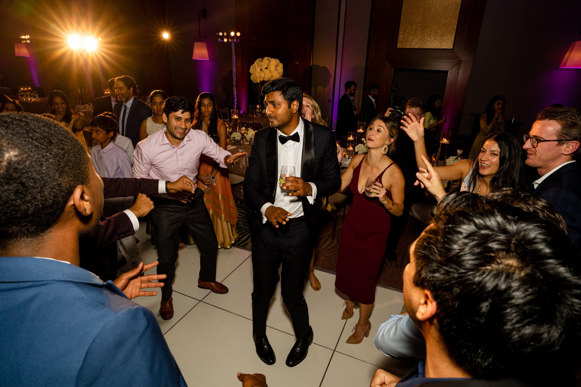 A group of people dancing energetically at a party with dim lighting. A man in a black tuxedo is in the center, holding a drink. Others around him are smiling and moving to the music. The background is softly lit, creating a festive atmosphere.