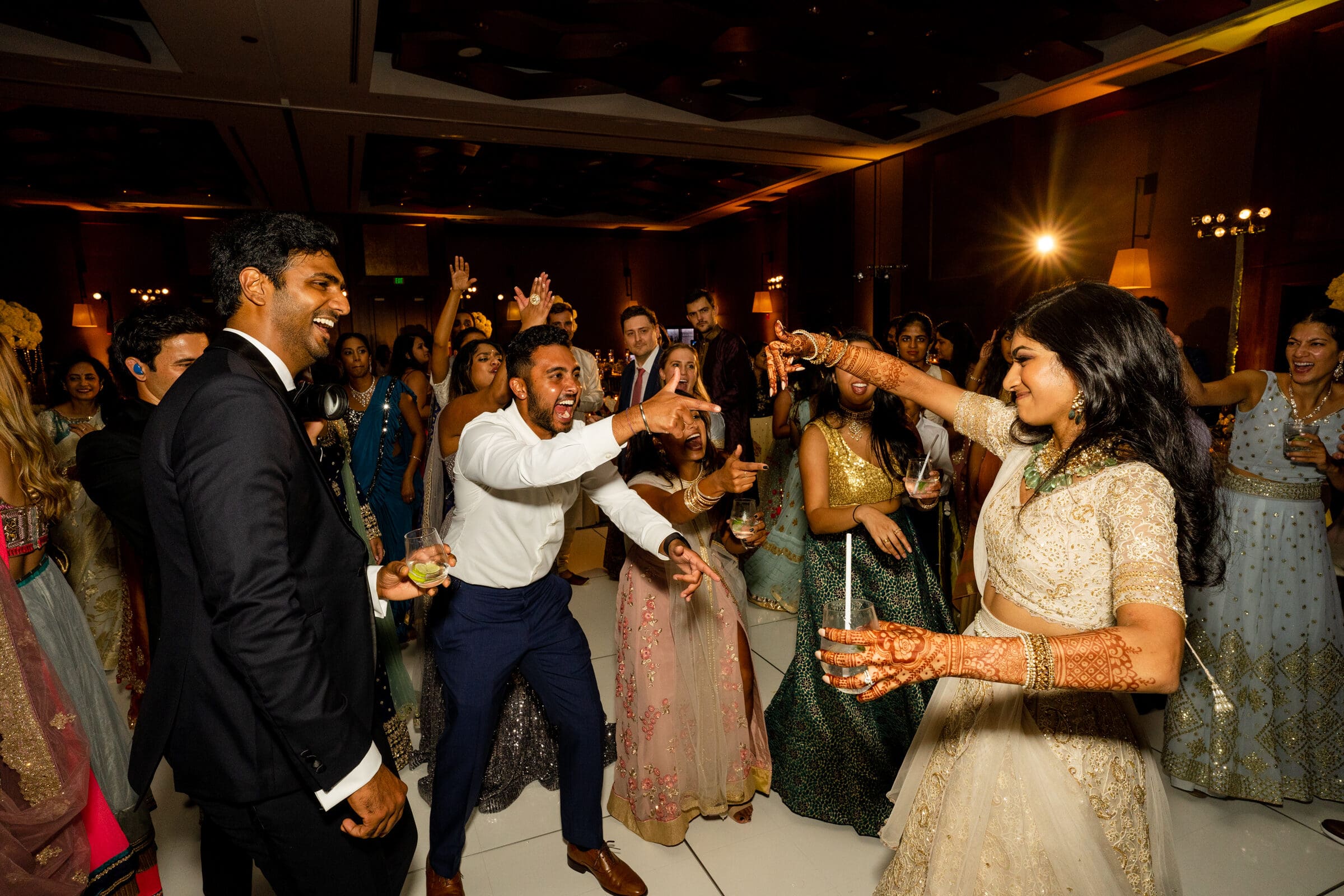 A lively dance floor with a group of people celebrating at a party. The focus is on a woman in a cream-colored outfit and a man in a white shirt enthusiastically dancing. Others in colorful attire are joyfully engaged in the background.