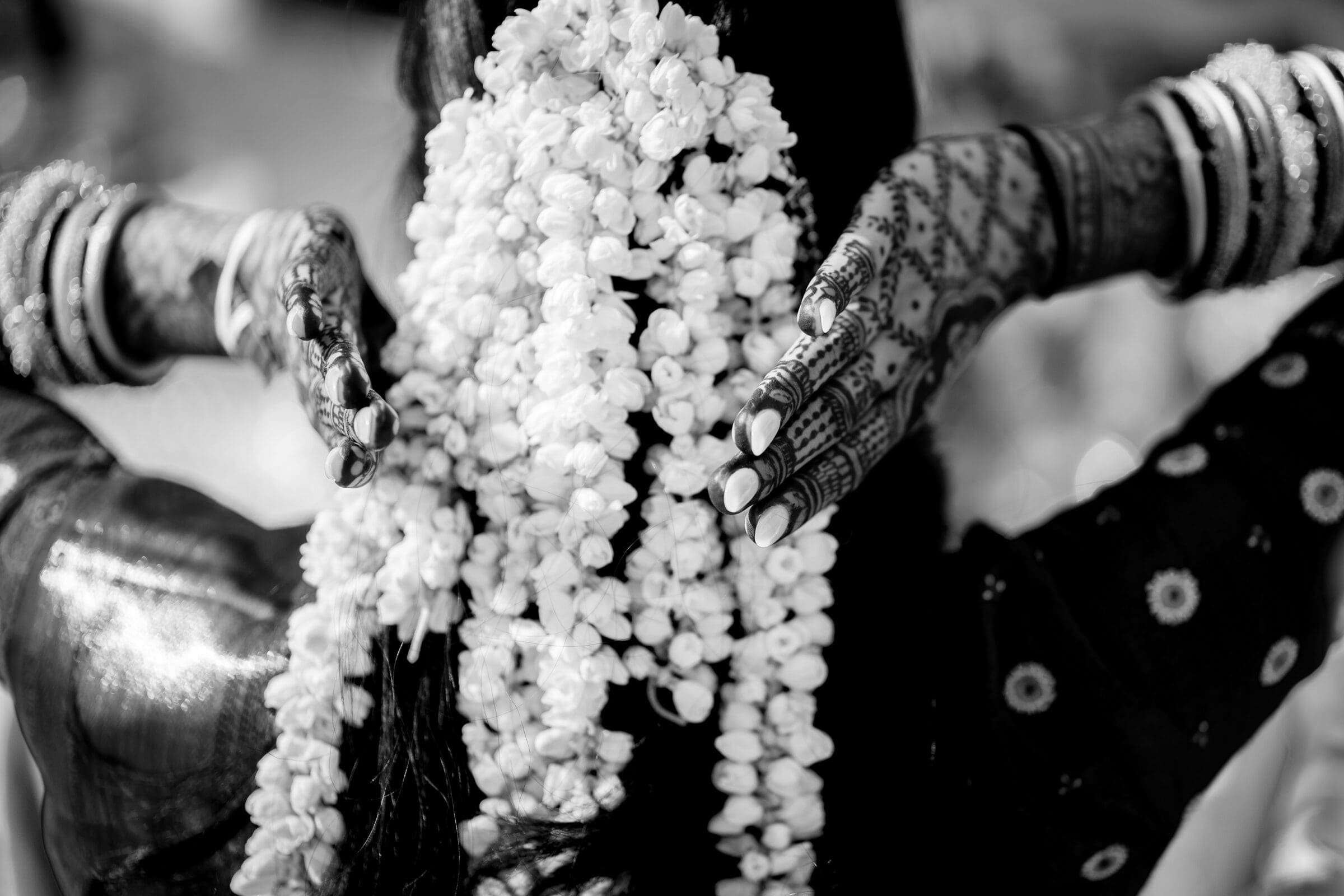 A person with intricate henna designs on their hands adjusts a garland of white flowers in their hair. They are wearing multiple bangles, and the focus is on the detailed patterns and textures, captured in black and white.