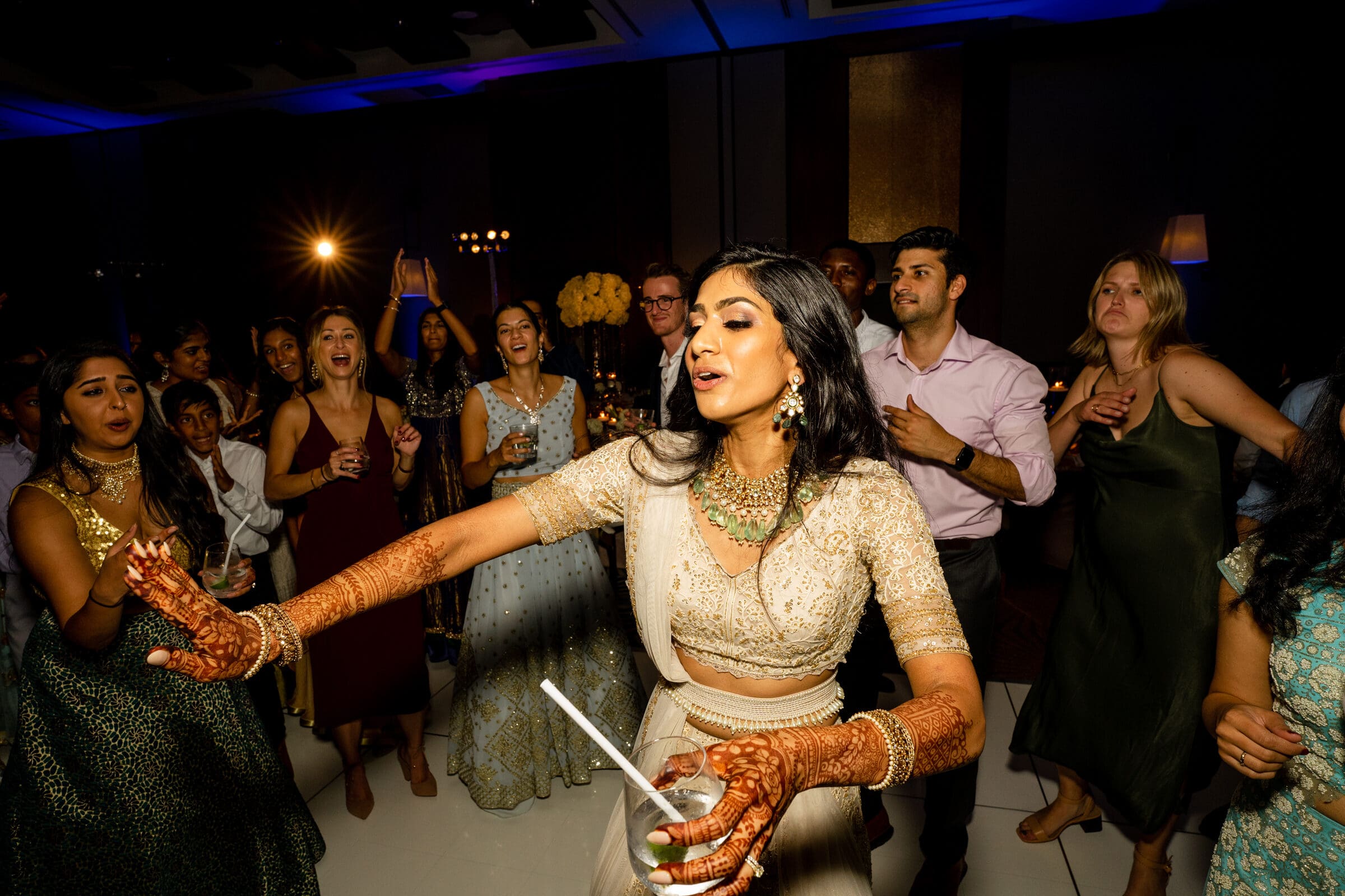 A woman in traditional Indian attire dances energetically on a dance floor, surrounded by people who are clapping and cheering. She has intricate henna designs on her arms and is holding a drink, with multicolored lights in the background.