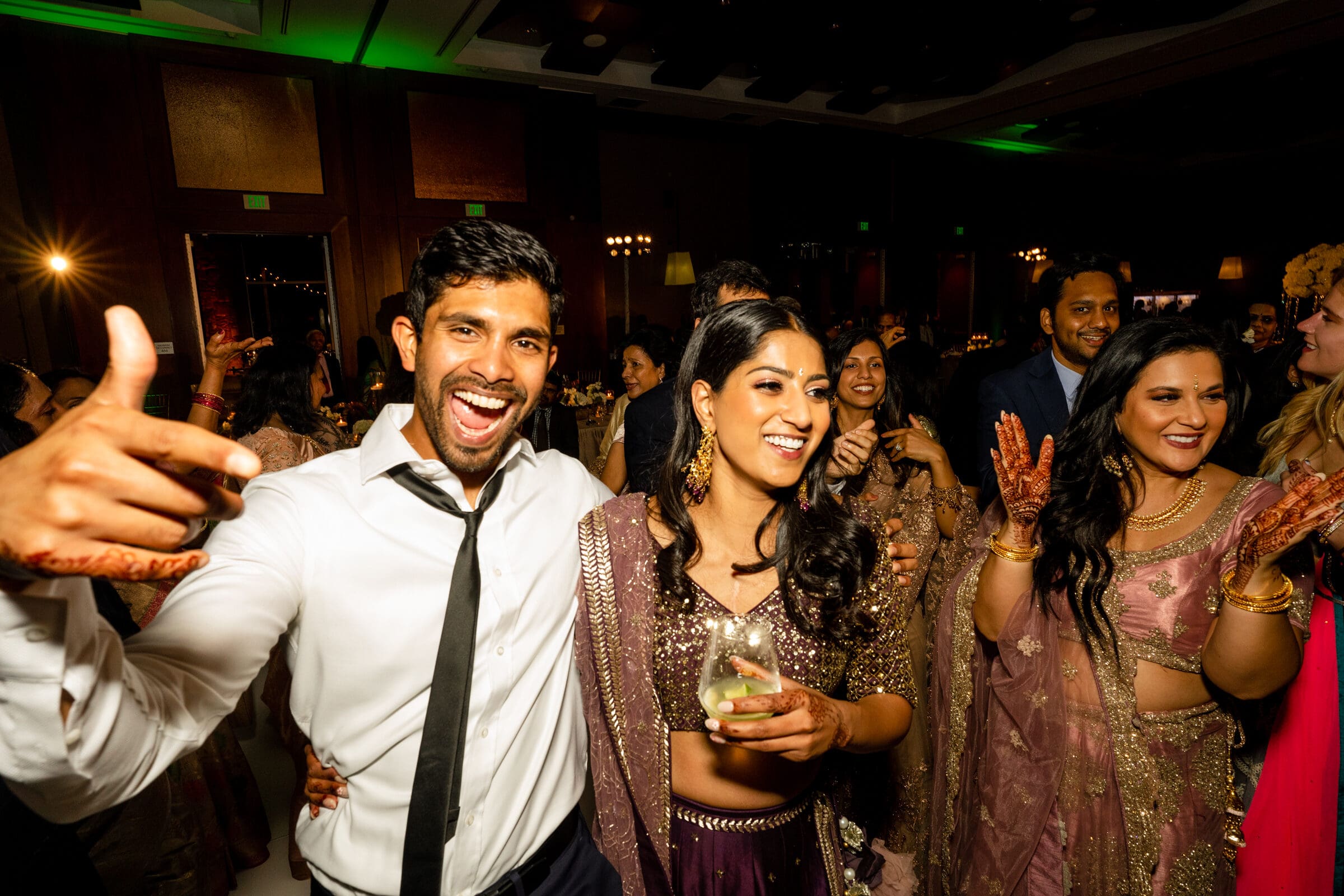 A group of people joyfully celebrating at a party. The man in the foreground is wearing a white shirt and tie, smiling and gesturing energetically. Next to him, a woman in a sequined outfit holds a drink and laughs. Others are dancing and enjoying the moment.