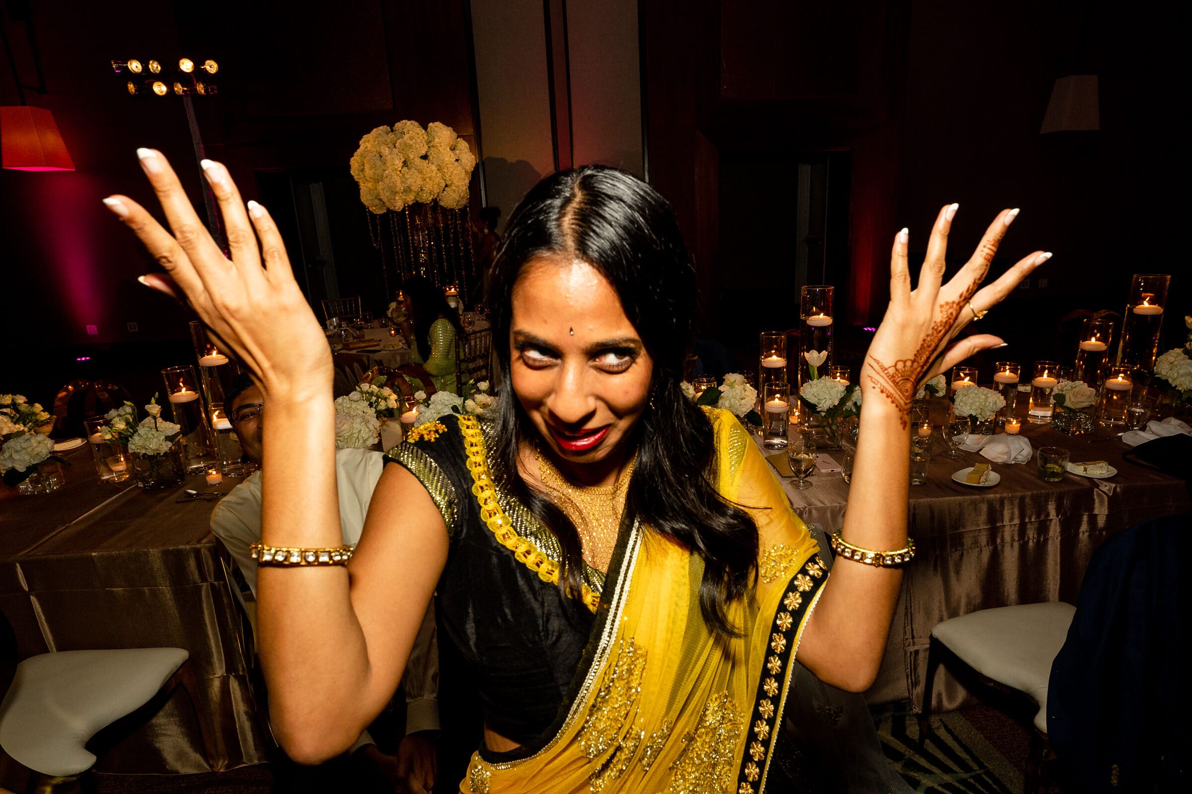 A woman in a black and yellow sari playfully gestures with her hands raised. She is smiling, with decorated arms and long dark hair. The background features a dimly lit event space with a table, floral arrangements, and candles.