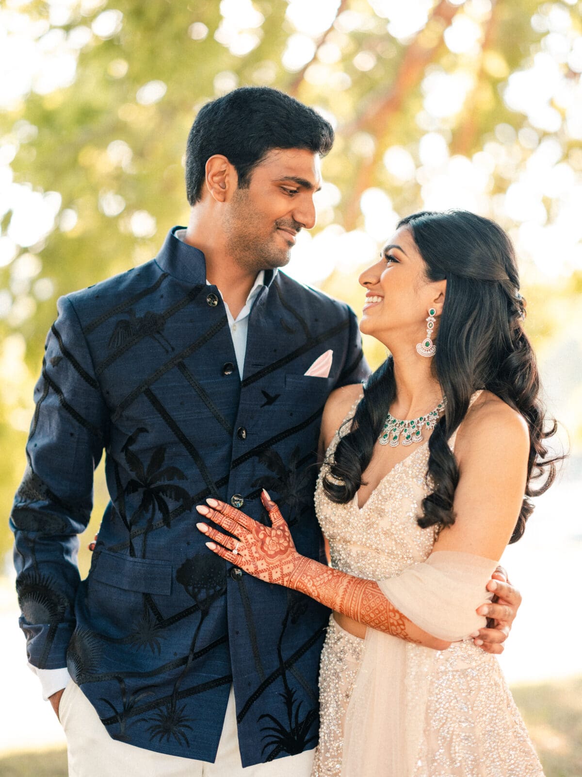 A couple dressed in elegant traditional attire, with the woman wearing intricate henna on her hands. They smile at each other against a blurred background of green foliage, creating a warm and joyful atmosphere.