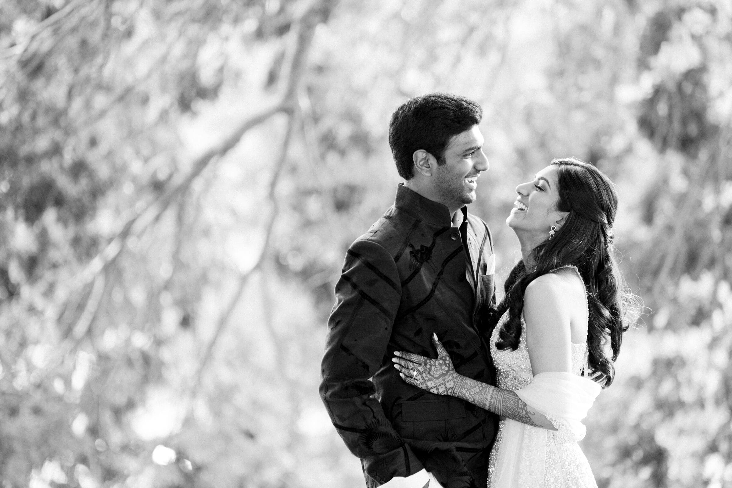 A black and white photo of a couple smiling at each other outdoors. The woman wears a dress with intricate henna on her hands, while the man wears a suit. They are embraced amid a softly blurred natural background.