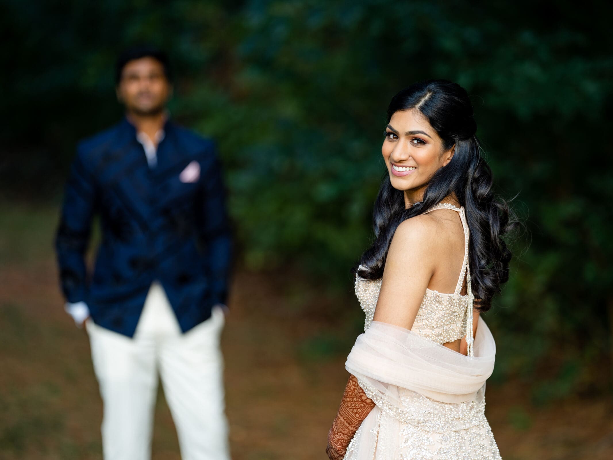 A woman in an elegant dress adorned with sequins smiles at the camera, standing in the foreground. A man in a dark blue suit stands behind her, slightly out of focus, set against a backdrop of lush greenery.