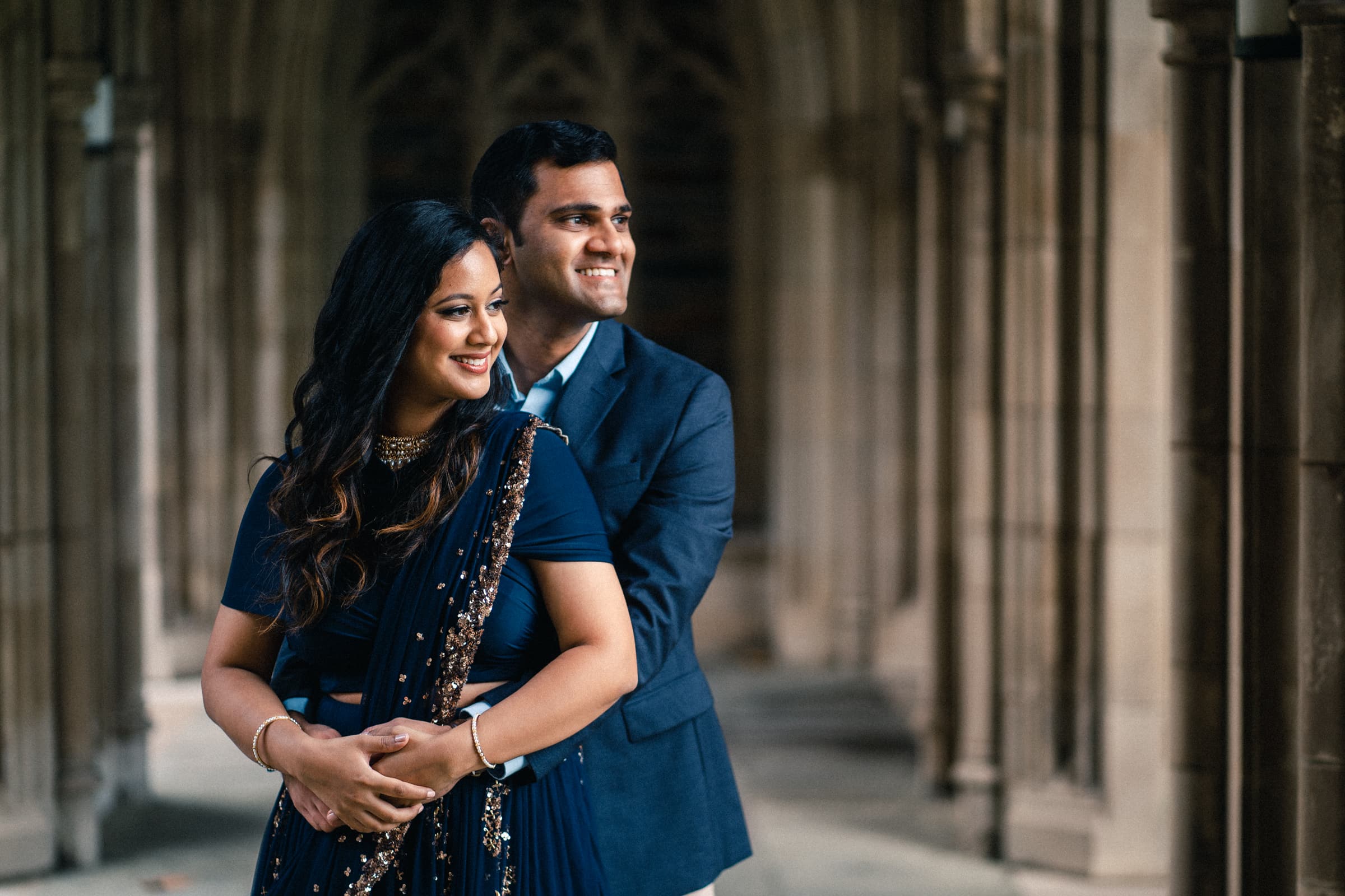 duke university engagement photos - shreena & kisan A couple stands in a stone hallway with arches. Both are smiling, looking off into the distance. The woman is wearing a dark blue saree with gold embellishments, and the man is dressed in a blue suit. They appear relaxed and happy.