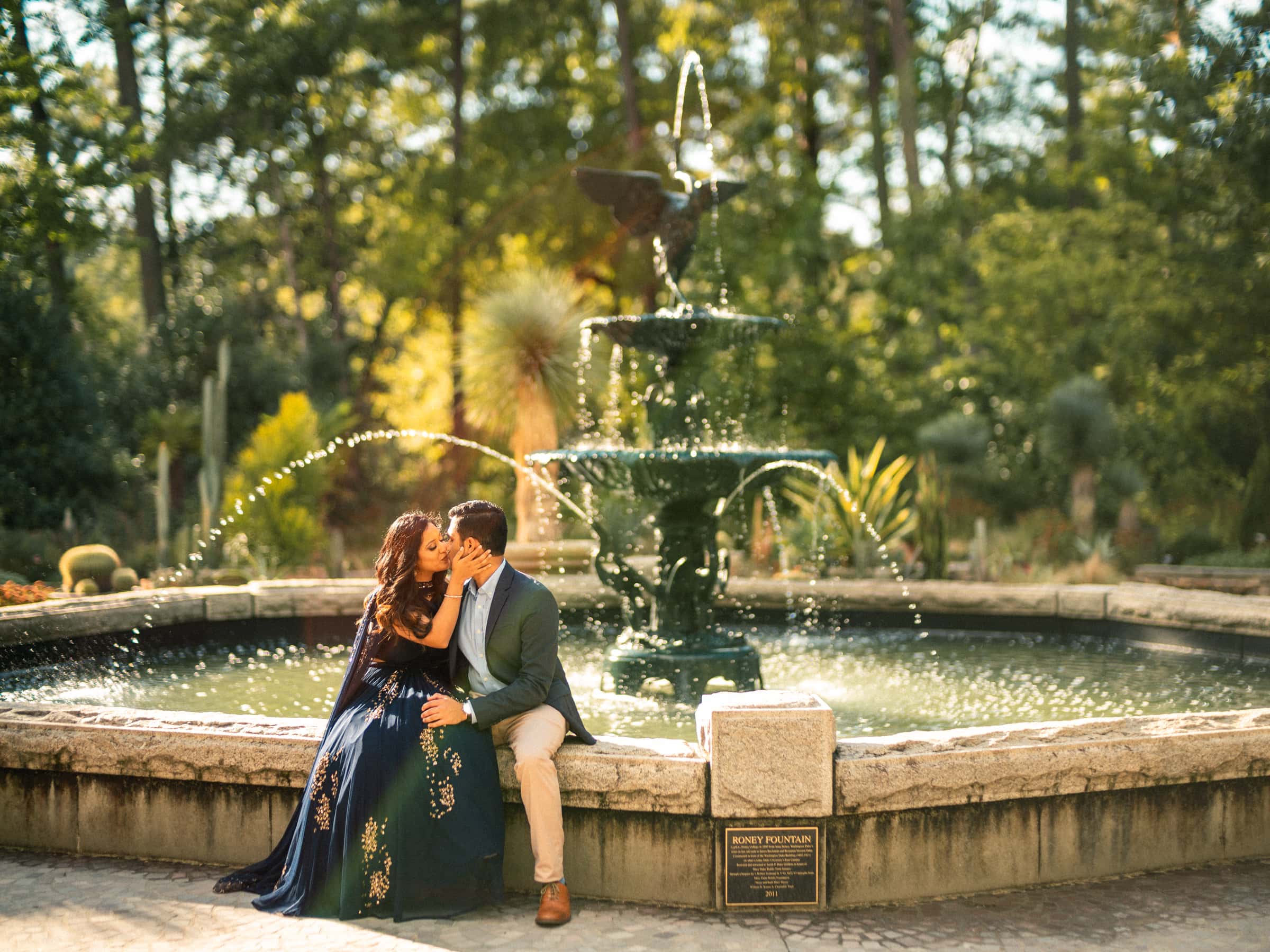 duke university engagement photos - shreena & kisan A couple sits by a large, ornate fountain in a lush garden. The woman in a navy dress and the man in a suit share a kiss while sunlight filters through the trees, creating a warm, romantic atmosphere.