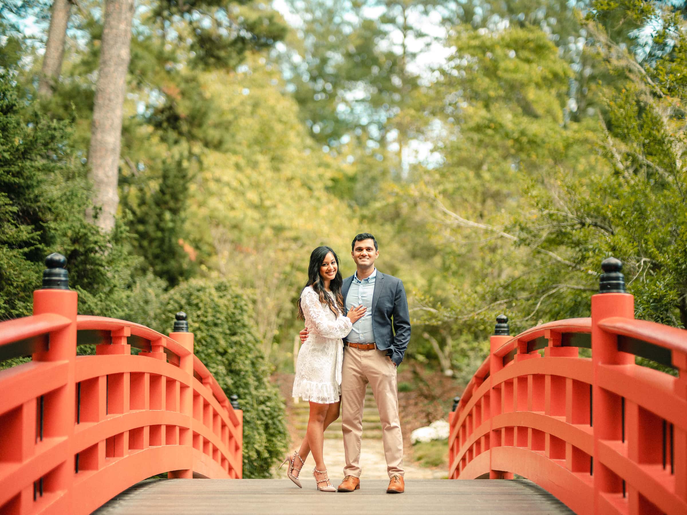 duke university engagement photos - shreena & kisan A couple stands on a red arched bridge surrounded by lush greenery. The woman is wearing a white dress, and the man is in a blue shirt with a blazer and beige pants. They are smiling with trees and foliage in the background.