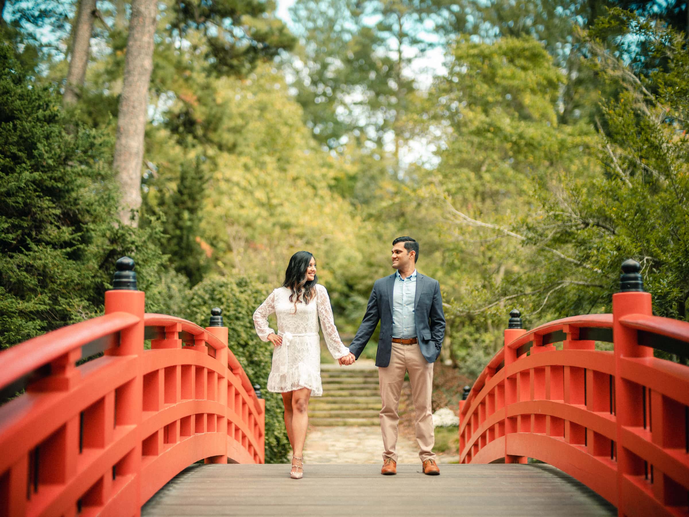 duke university engagement photos - shreena & kisan A couple walks hand in hand on a red arched bridge in a lush garden. The woman wears a lace dress, and the man is in a suit. They are smiling at each other, surrounded by green trees and foliage.