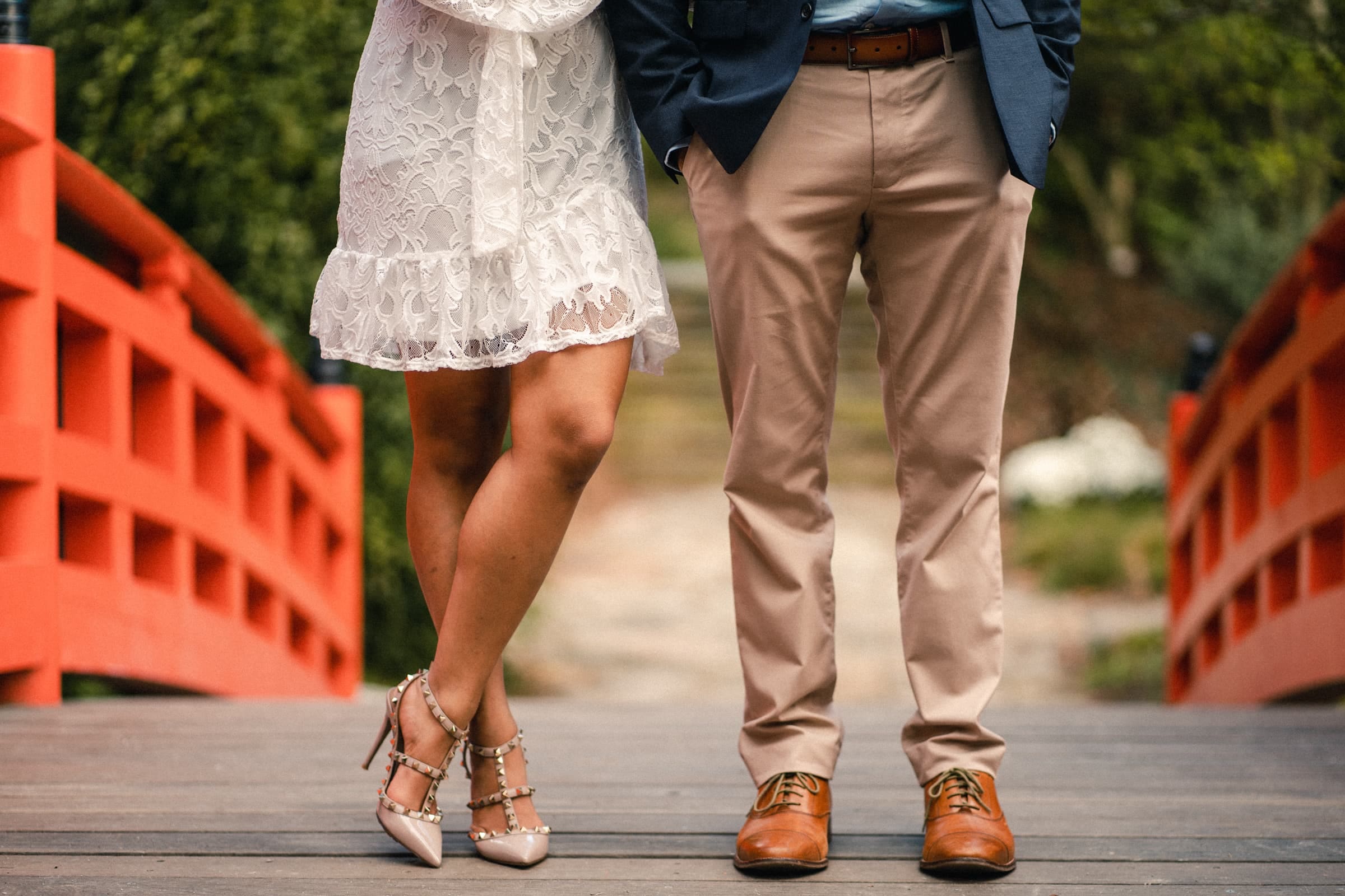 duke university engagement photos - shreena & kisan A couple stands side by side on a wooden bridge with a red railing. The woman is wearing a white lace dress and studded heels, while the man is in a navy blazer, beige pants, and brown shoes. The background is a blurred, lush green setting.