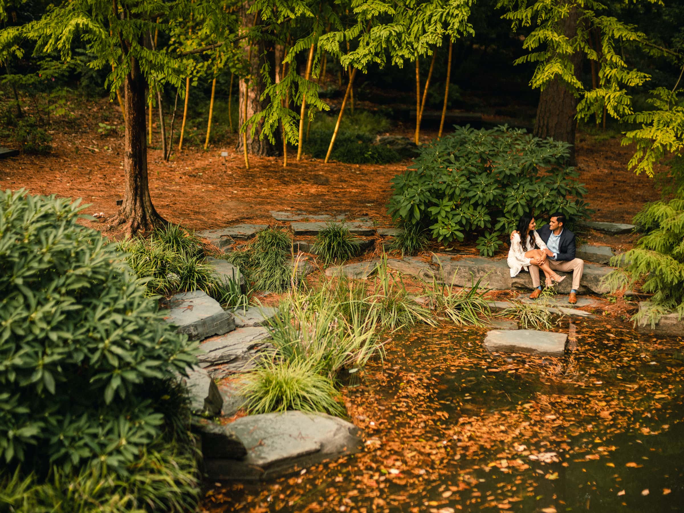duke university engagement photos - shreena & kisan A couple sits on stone steps beside a tranquil pond surrounded by lush greenery and tall trees. The ground is covered with fallen leaves, adding a touch of autumn to the serene natural setting.