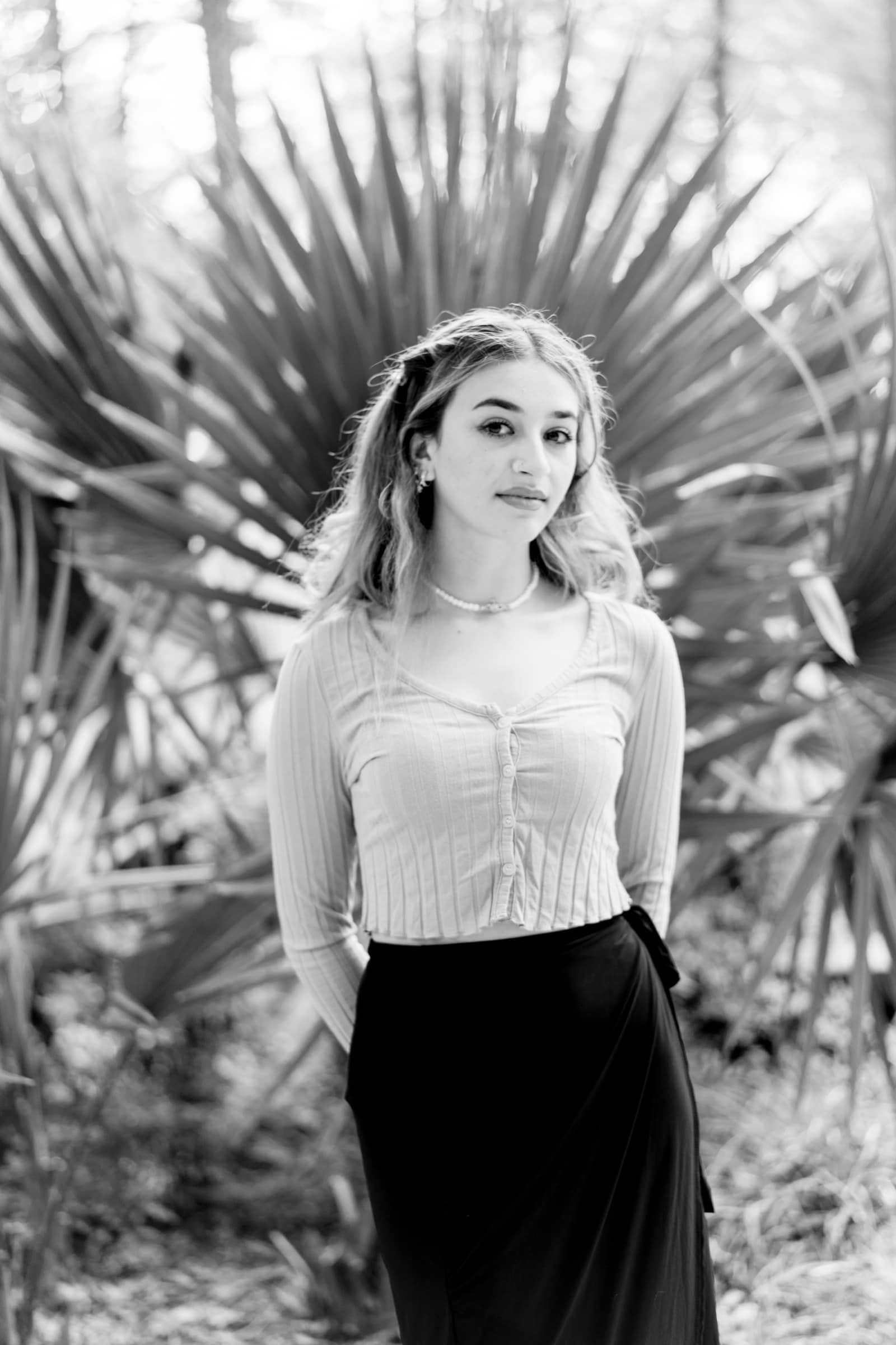 durham portrait photographers - sophia's senior session A woman stands gracefully in front of large, spiky leaves. Captured by a Durham portrait photographer, she wears a light-colored, long-sleeved top and a dark skirt. The image in black and white exudes elegance as her hands rest discreetly behind her back.