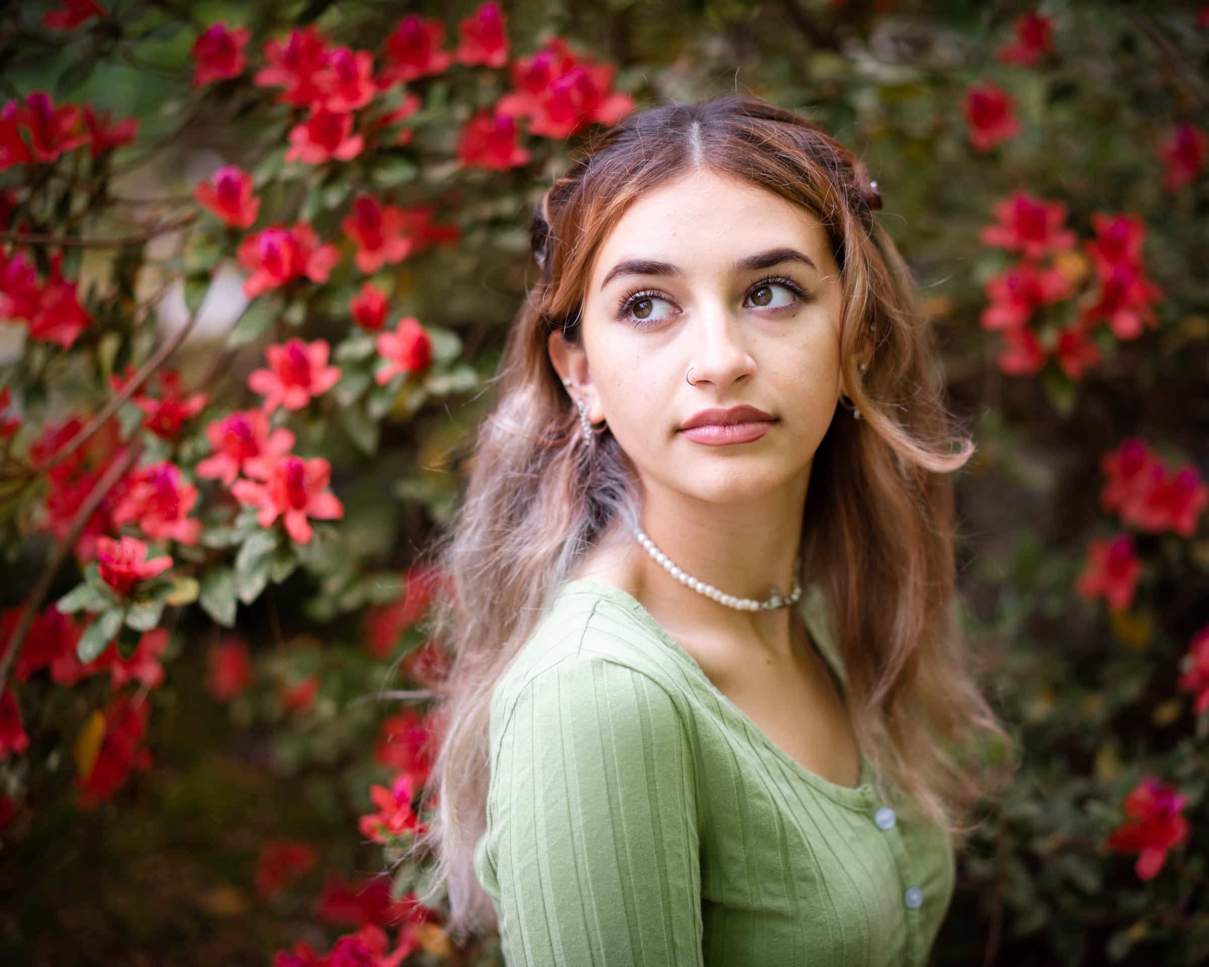 durham portrait photographers - sophia's senior session A young woman with long hair stands in front of vibrant red flowers, captured by a Durham portrait photographer. She wears a light green top and a pearl necklace, gazing thoughtfully to the side with a soft and serene expression.