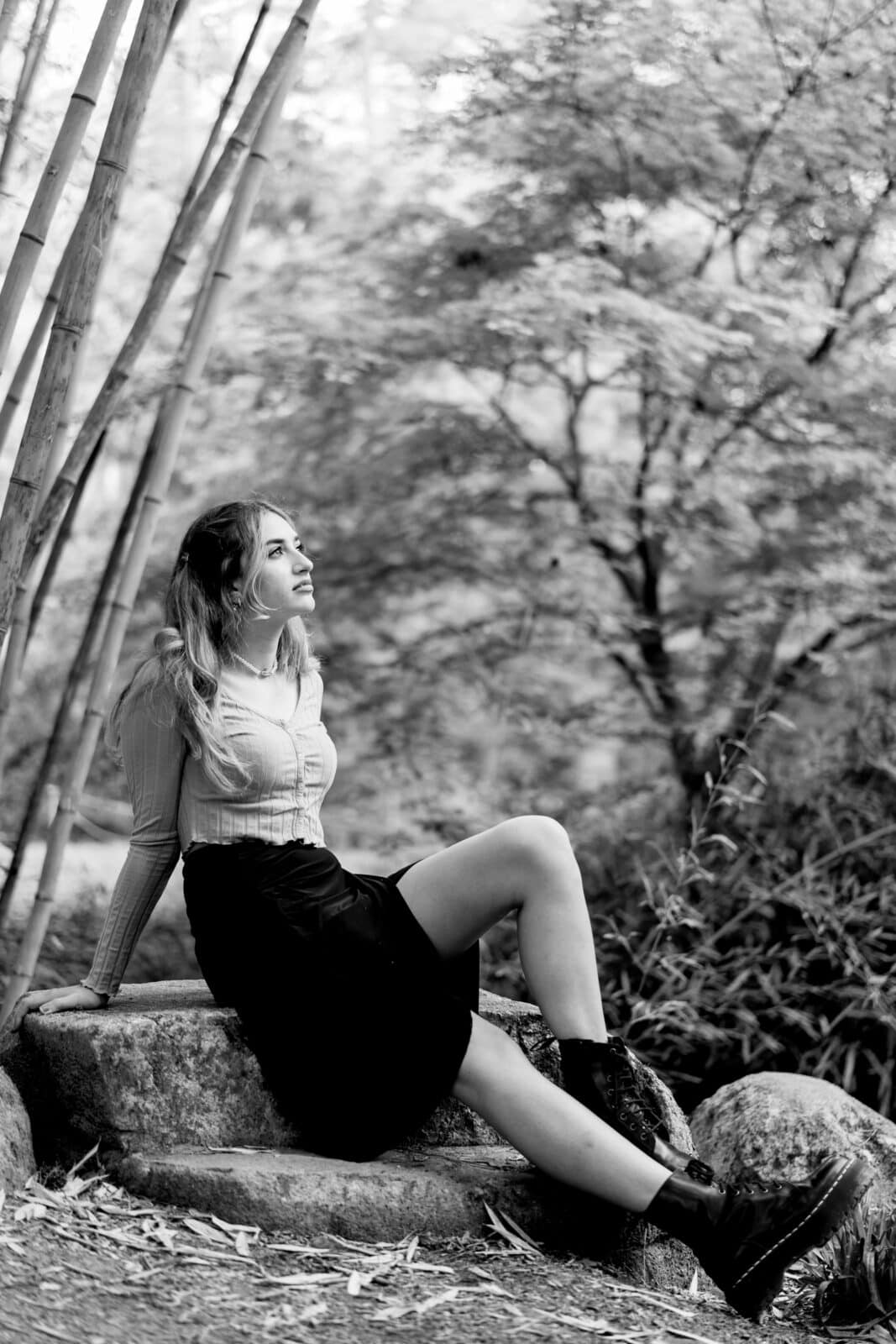 durham portrait photographers - sophia's senior session A woman sits on stone steps surrounded by bamboo and lush foliage. She is dressed in a light blouse, dark skirt, and boots, gazing upward with a thoughtful expression. Captured by a Durham portrait photographer, the image is beautifully rendered in black and white.