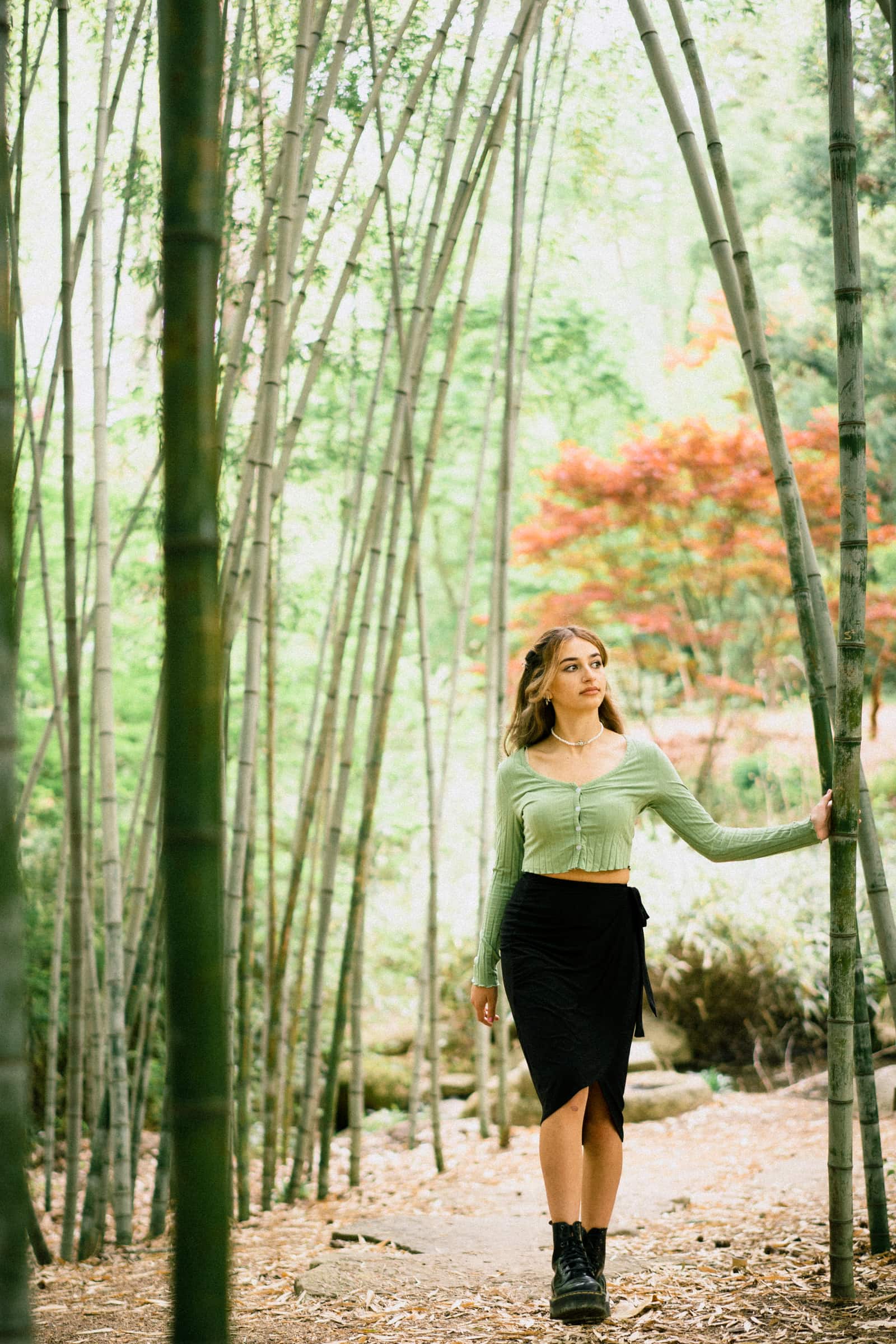 durham portrait photographers - sophia's senior session A woman in a green top and black skirt strolls through a bamboo forest, gently touching a stalk while gazing thoughtfully into the distance. Captured with the artistry of a Durham portrait photographer, vibrant foliage adds to the scene’s enchanting allure.