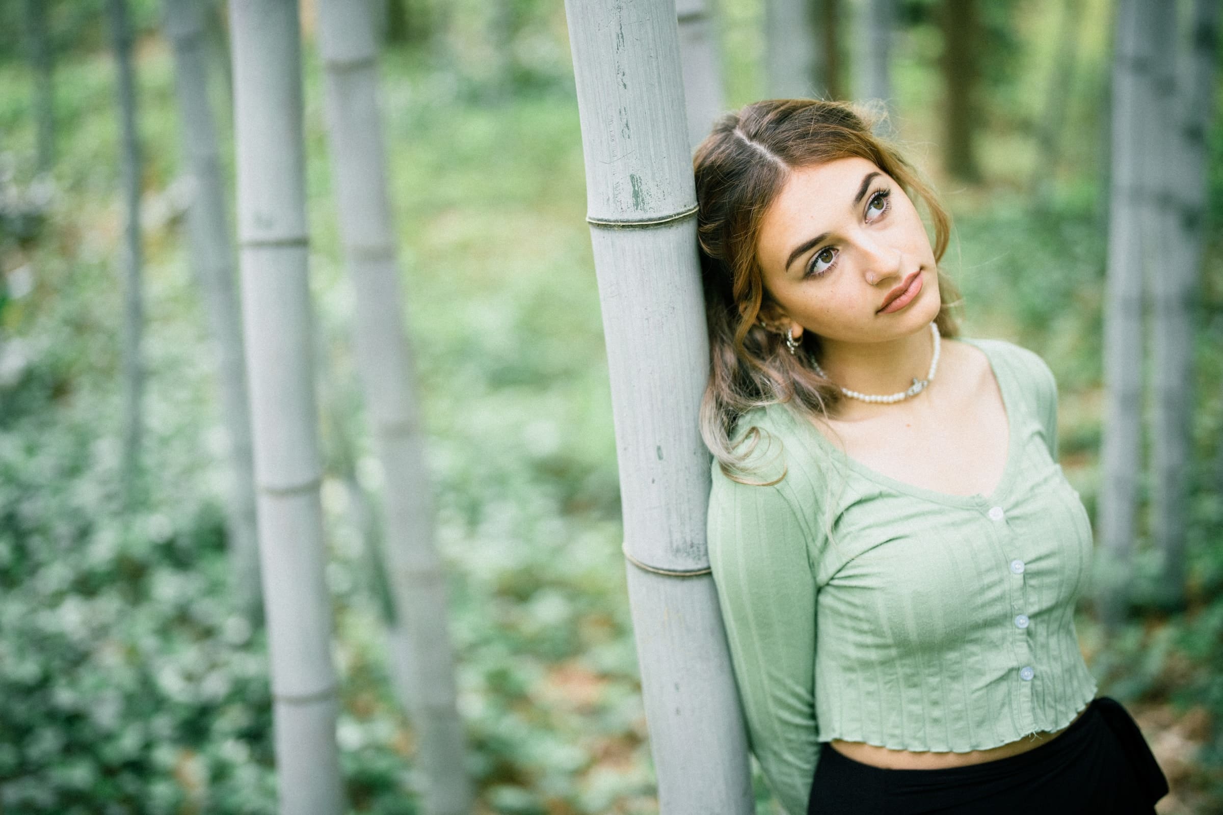 durham portrait photographers - sophia's senior session A young woman in a green top leans against a bamboo tree, gazing thoughtfully upward. Captured by a Durham portrait photographer, she is surrounded by a lush, green forest environment.