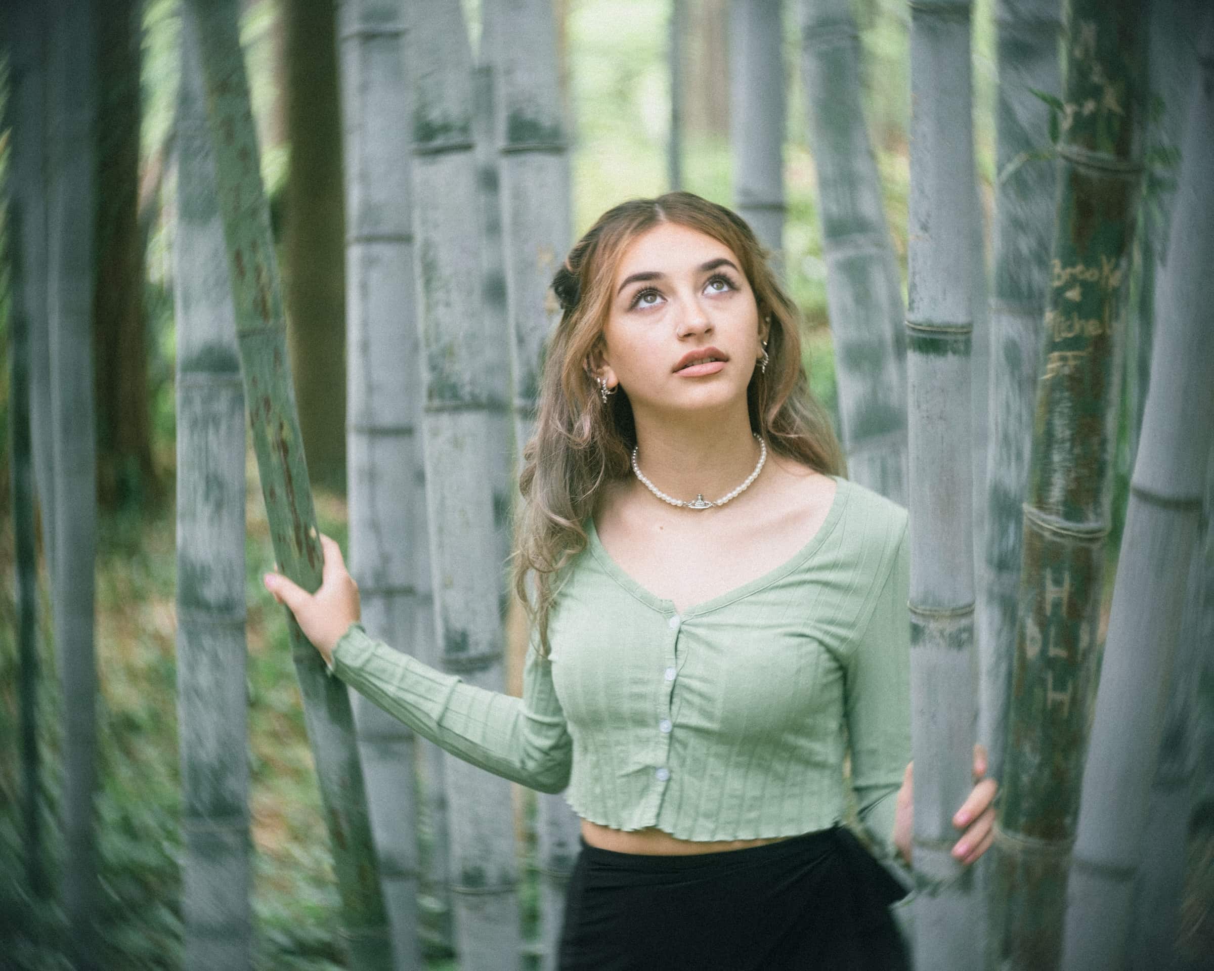 durham portrait photographers - sophia's senior session A woman in a green top stands among tall bamboo stalks, gazing upwards with a thoughtful expression. Captured by a skilled Durham portrait photographer, the setting is lush and serene, with soft, natural light illuminating the scene.