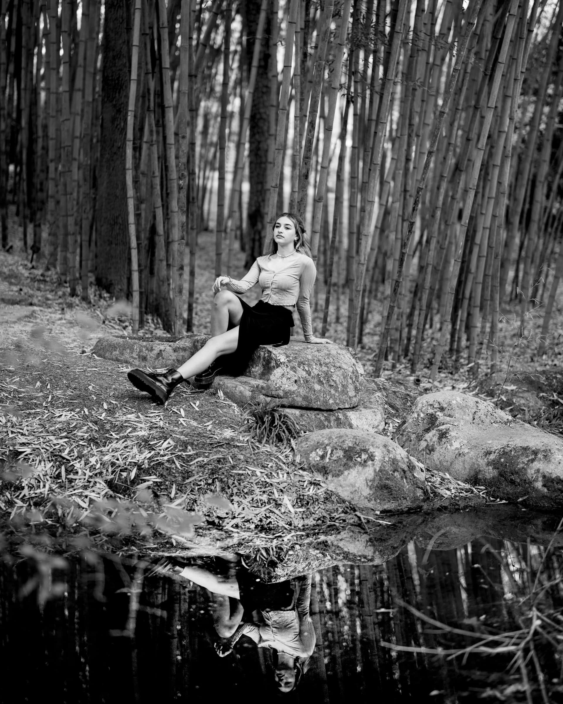 durham portrait photographers - sophia's senior session A woman sits on a rock beside a pond, surrounded by tall bamboo trees, captured by a Durham portrait photographer. She wears a light top and dark skirt, her reflection mirroring in the water. The scene in black and white evokes calmness and serenity.
