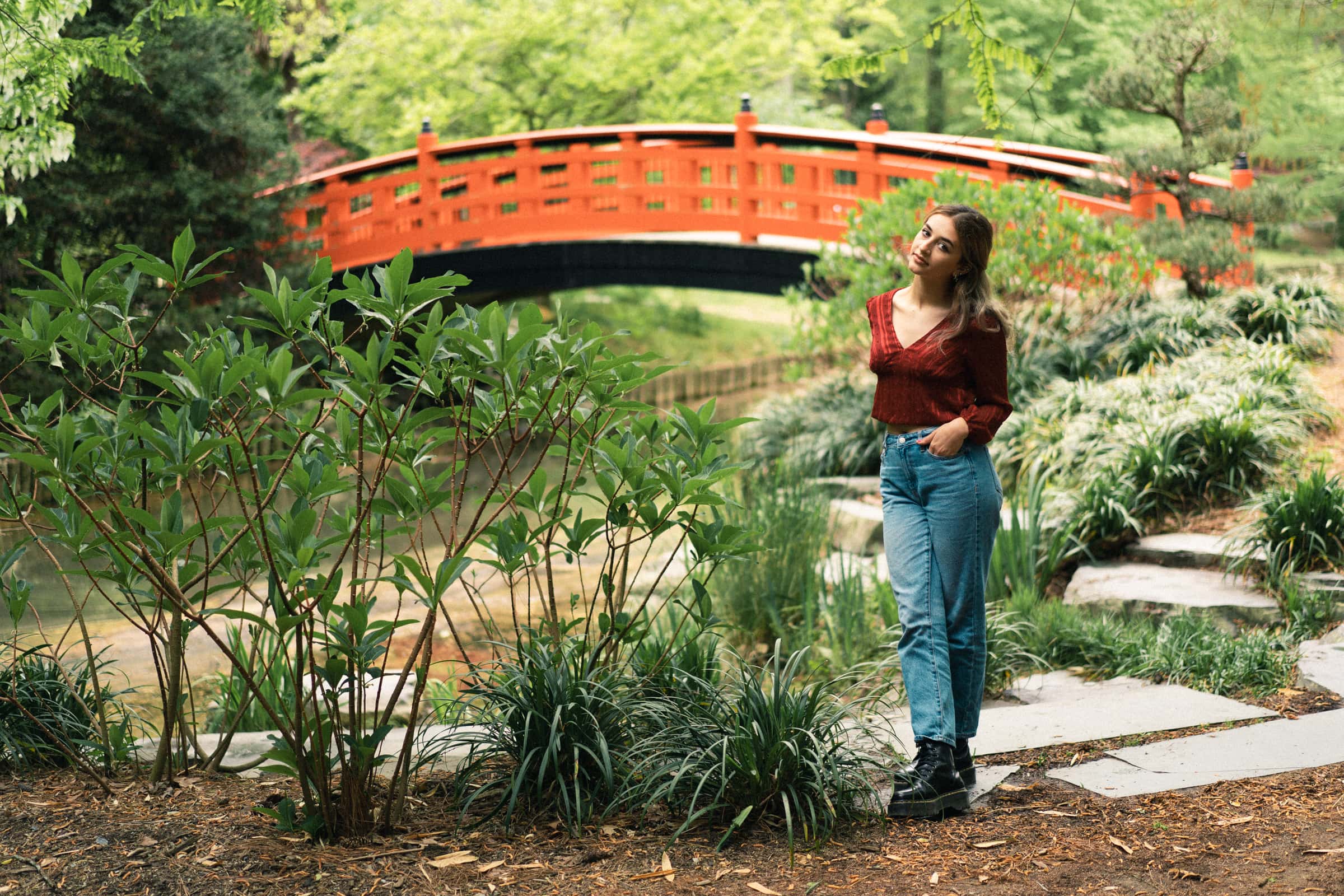 durham portrait photographers - sophia's senior session A woman stands on a garden path, surrounded by lush greenery and captured by a Durham portrait photographer. She wears a red blouse and blue jeans. In the background, a vibrant red arched bridge is beautifully set against trees and plants.