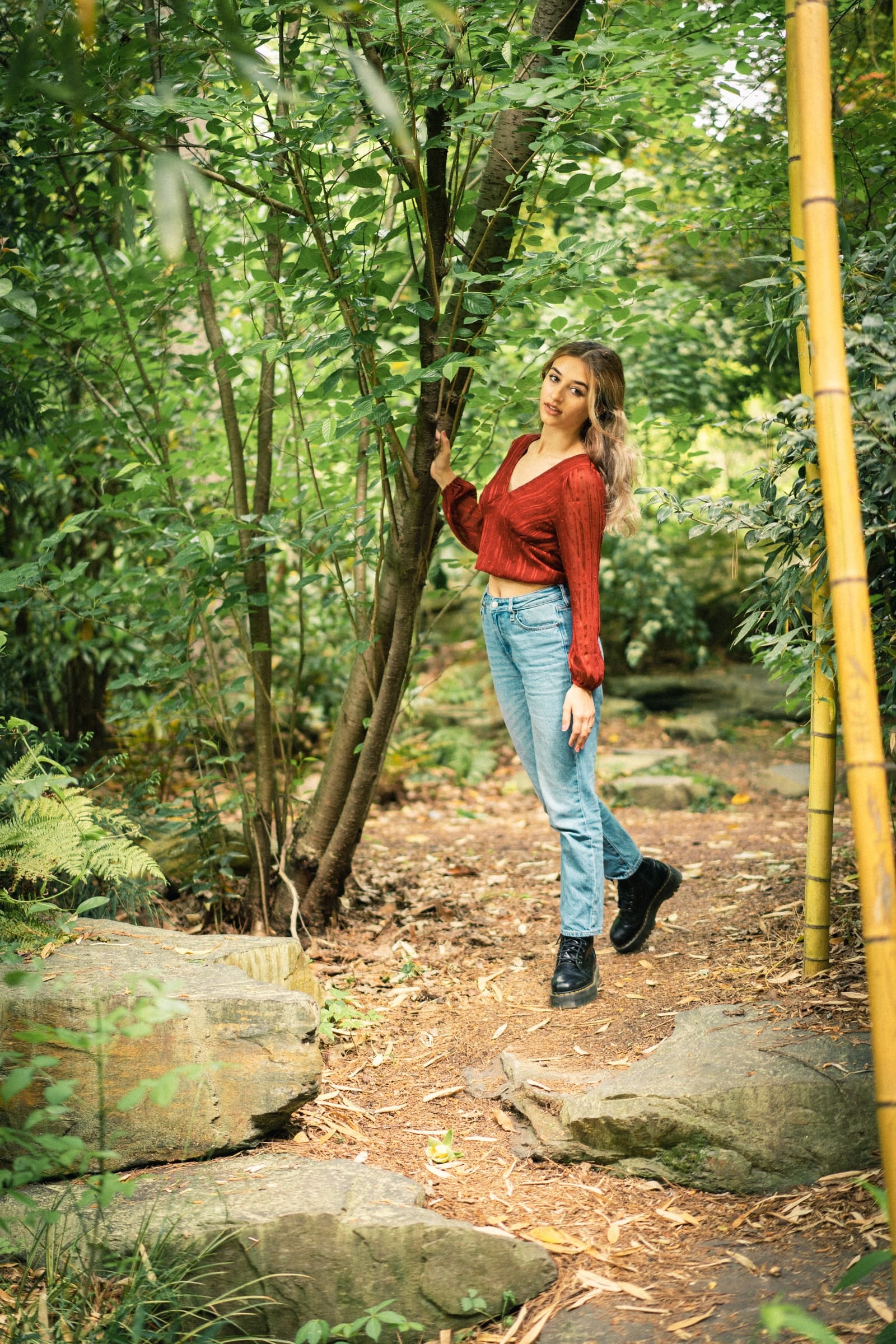 durham portrait photographers - sophia's senior session A woman in a red blouse and jeans stands on a forest path surrounded by green foliage and rocks. Captured with the eye of a Durham portrait photographer, she leans slightly against a tree with one hand, looking toward the camera. The setting is lush and serene.