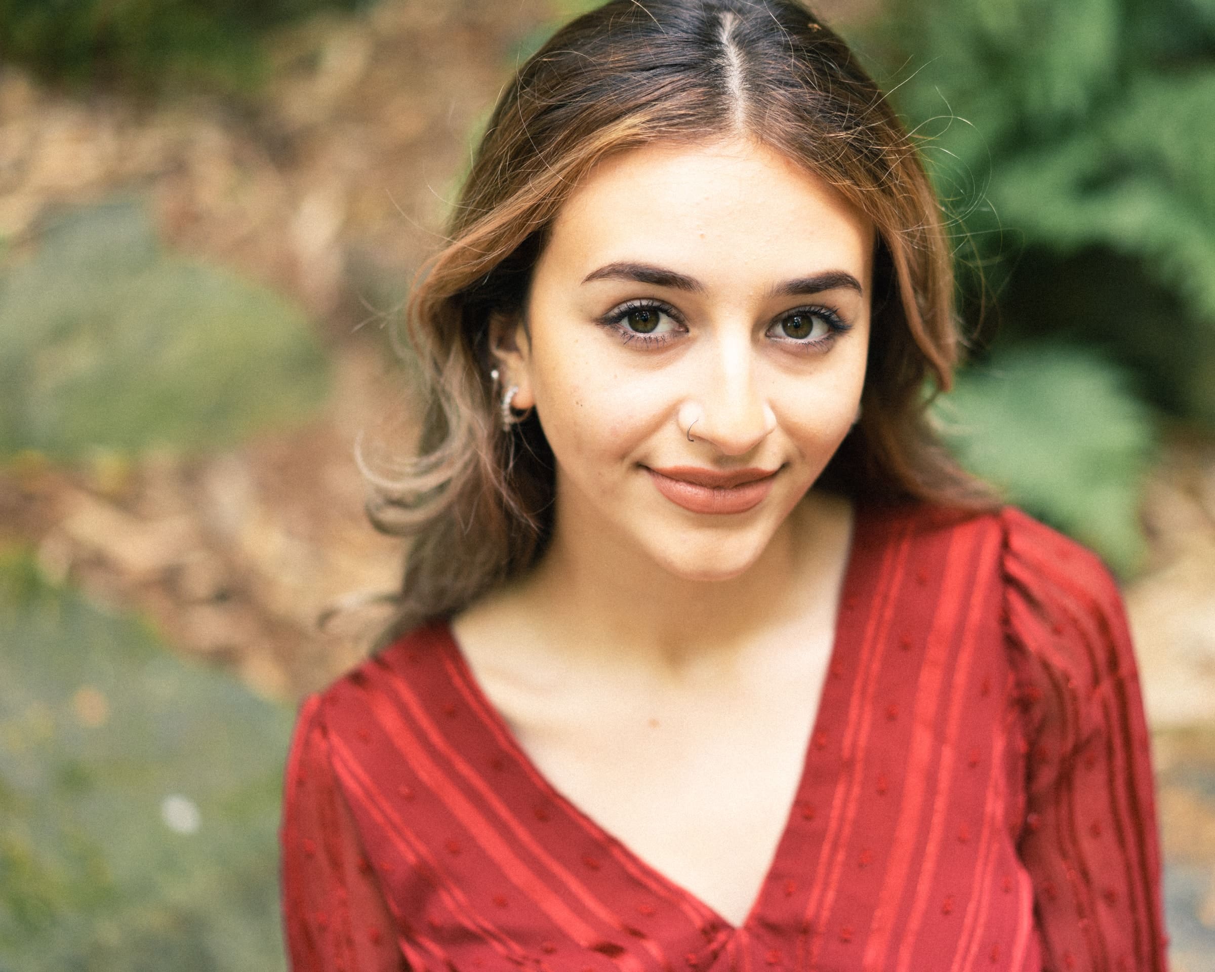 durham portrait photographers - sophia's senior session A young woman with long, wavy hair and a red blouse stands outdoors, captured by a talented Durham portrait photographer. She smiles gently at the camera, framed beautifully against a backdrop of blurred greenery.
