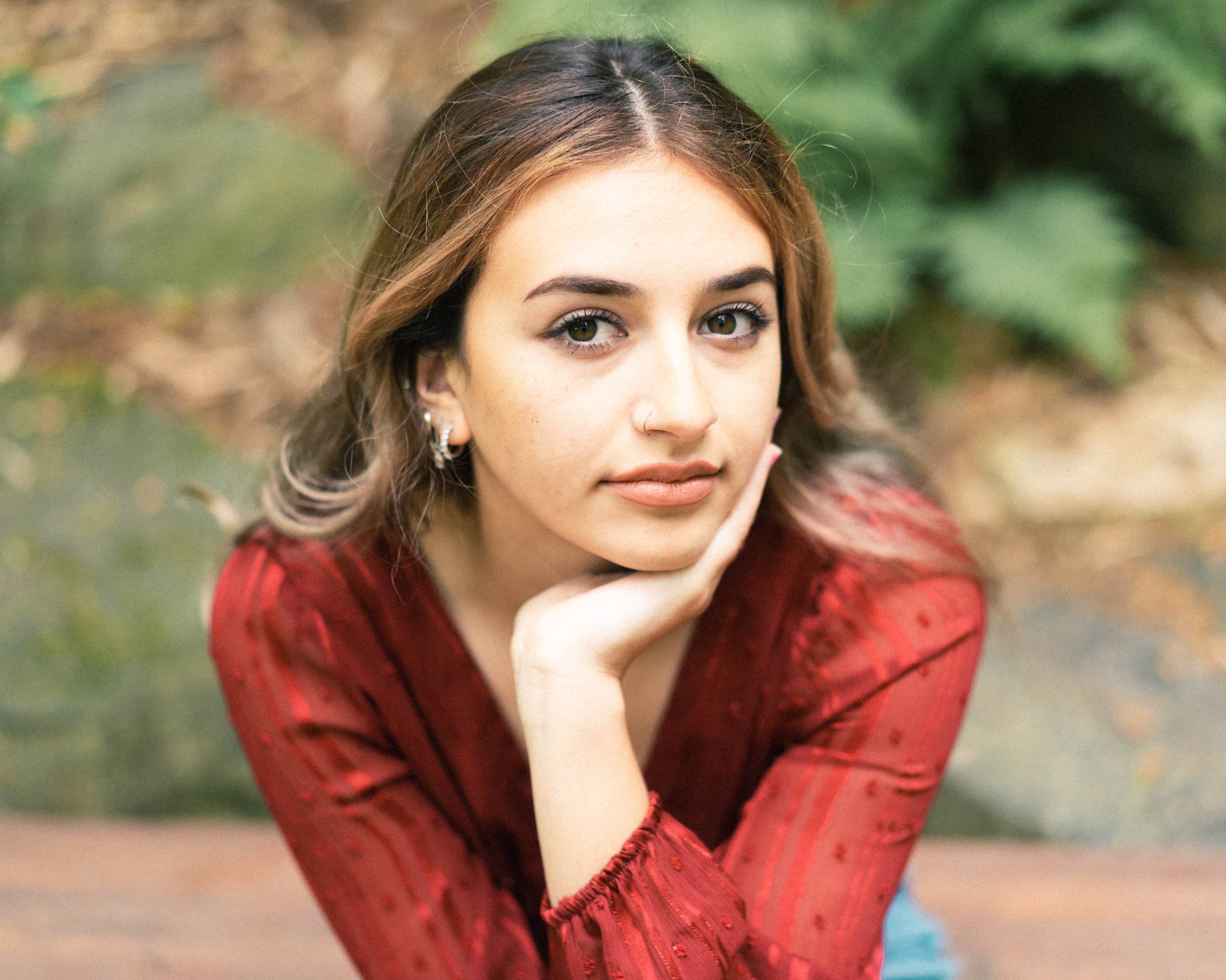 A person with long hair in a red top rests their chin on their hand, gazing thoughtfully at the camera. Captured by a Durham portrait photographer, the scene features a natural backdrop of green foliage and rocks.