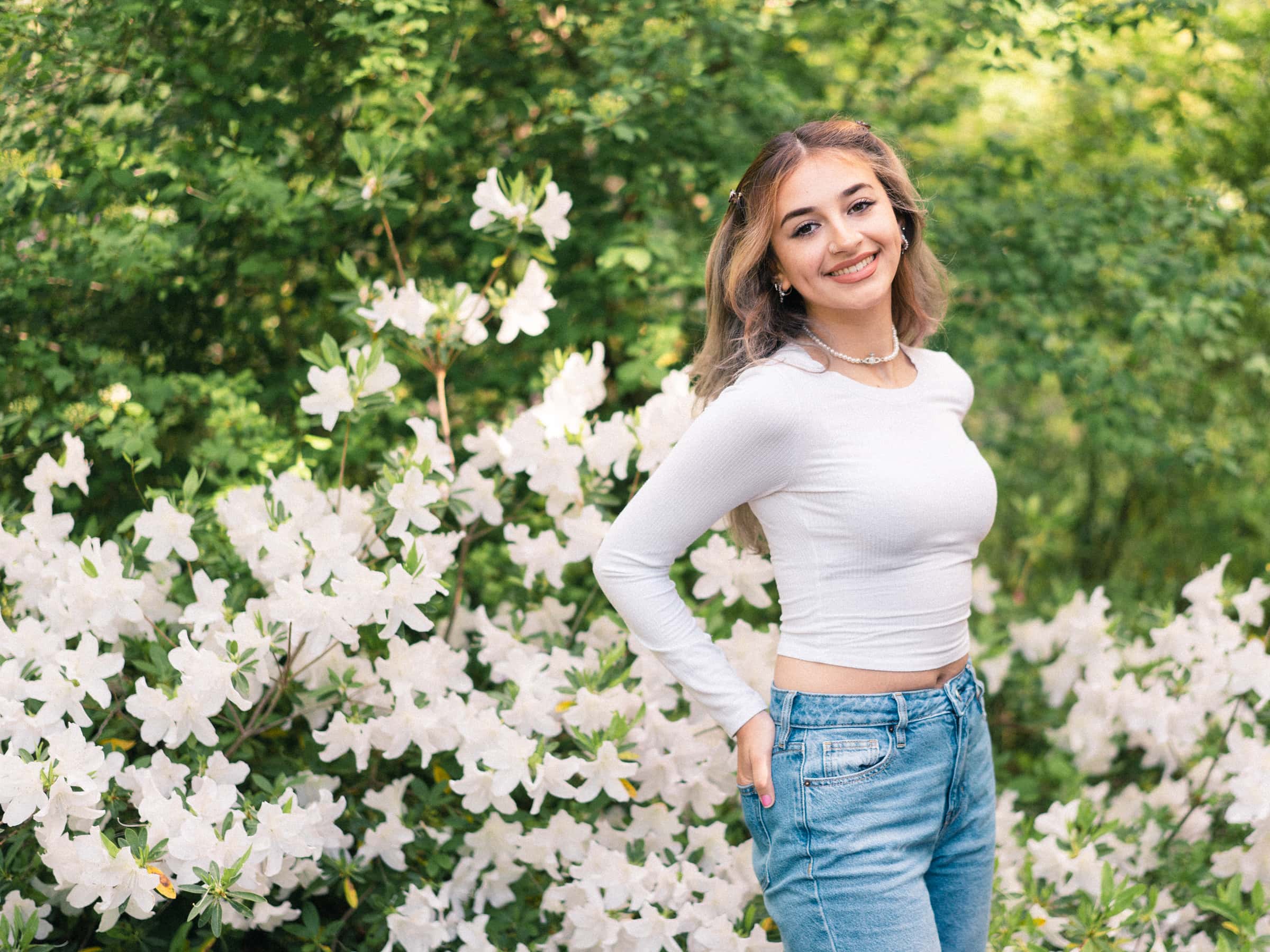 durham portrait photographers - sophia's senior session A person with long hair, wearing a white long-sleeve top and jeans, smiles while standing in front of white flowering bushes. The scene, reminiscent of a Durham portrait photographer's touch, is set against a lush backdrop of greenery.